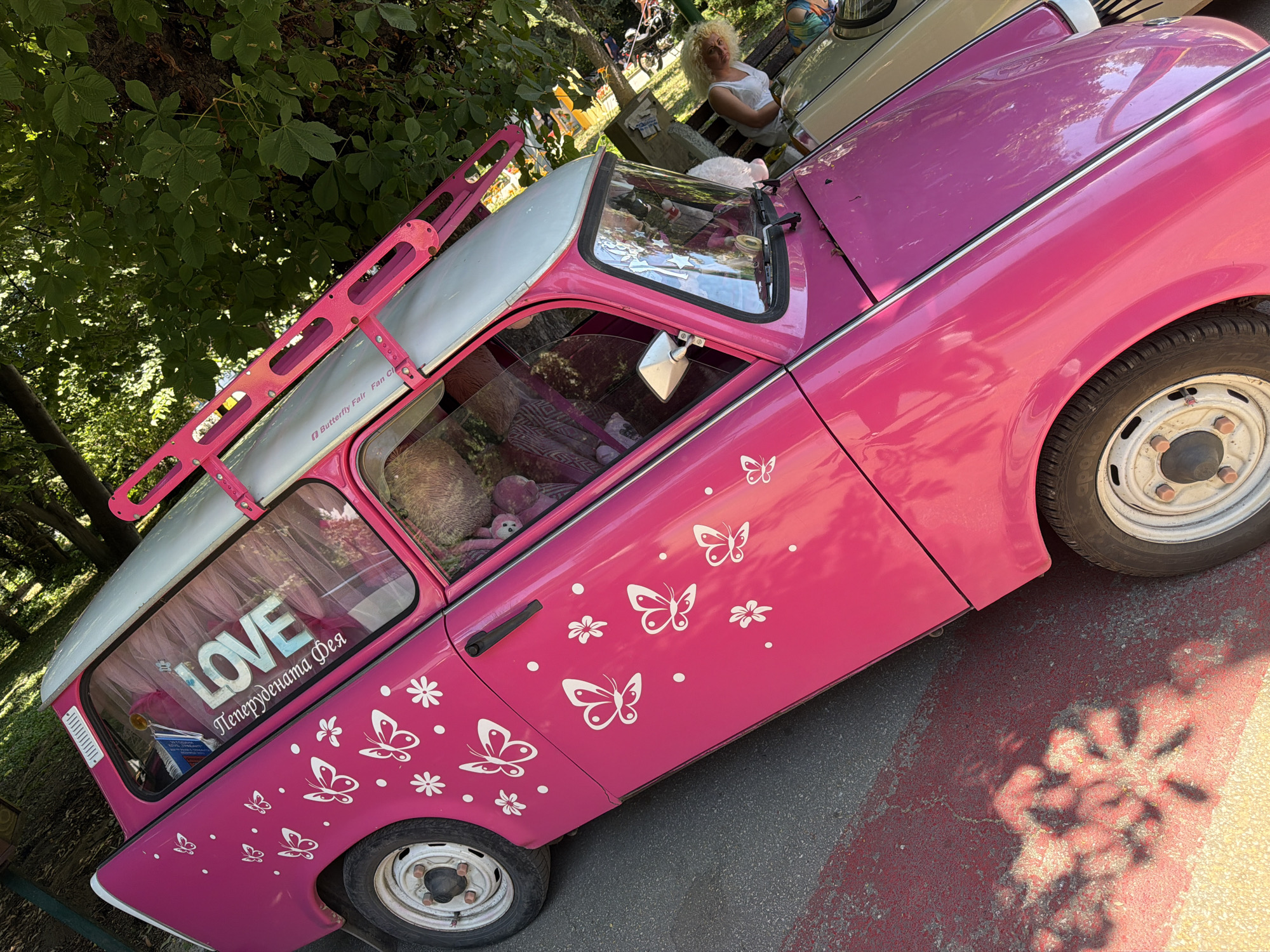 A bright pink Trabant car, adorned with white butterfly stickers and the word "LOVE" in large letters on its rear window, is parked on a street in Pazardzhik, Bulgaria. The car has a silver roof and a pink roof rack. In the background, there are trees with green foliage, and a woman with blonde, curly hair wearing a white top can be seen. The car is positioned on a paved surface with red and yellow markings.