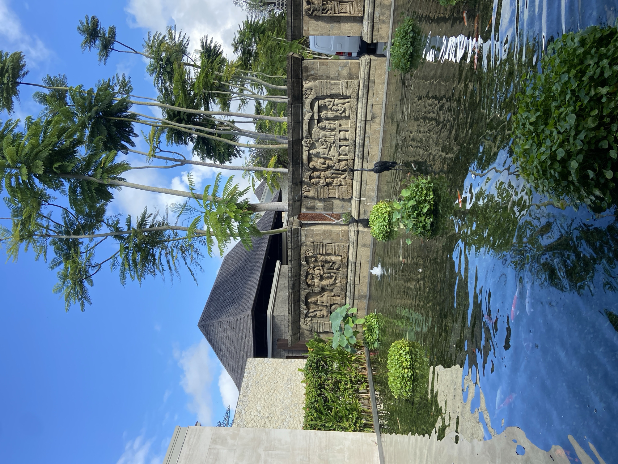 The image shows a tranquil scene with a pond in Kuta Selatan, Indonesia. Palm trees line the left side of the frame, contrasting against a blue sky with scattered clouds. An ornate stone wall with carved figures borders the pond. Reflected in the water are the sky, trees, and carvings. Plants and vegetation are strategically placed around the pond. A black bird stands near the edge of the water. Koi fish are visible swimming in the pond. Part of a building with a dark roof and a light-colored stone facade is visible behind the wall. A vehicle is partially visible behind the wall.
