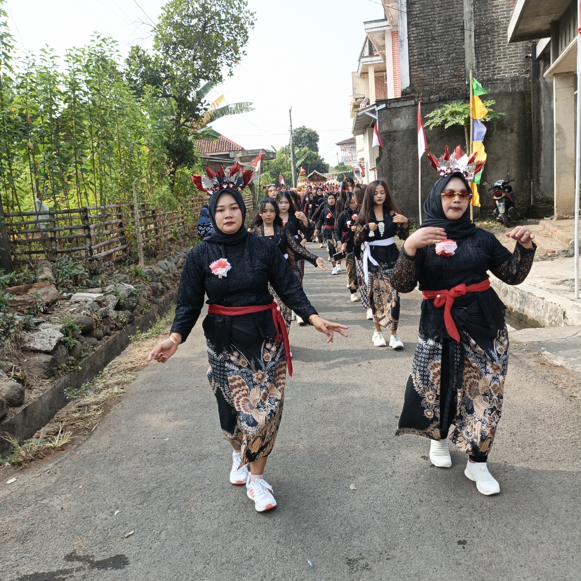In Tanahbaya, Indonesia, a procession of young women walks down a paved road lined with trees and bamboo fencing. The women are dressed in traditional black lace blouses and batik skirts, cinched with red sashes. Many wear elaborate headpieces adorned with red and silver feathers. Some women have white flowers pinned to their blouses. The procession appears to be a cultural performance or celebration. Flags, including Indonesian flags, are visible on poles in the background, suggesting a national holiday or event. The road is bordered by a concrete drainage ditch and rocky terrain on the left, and buildings and a parked motorcycle on the right. The sky is bright and clear.