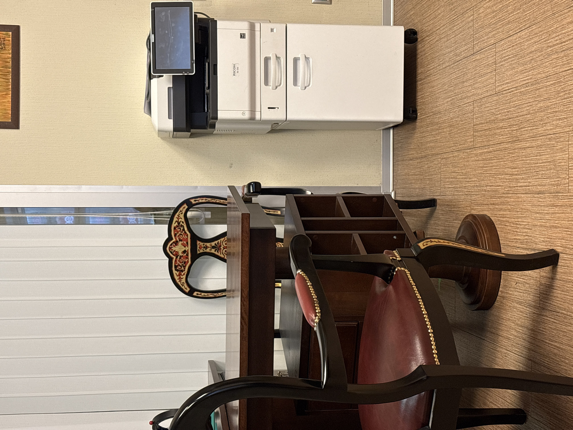 The image shows a room setting with a wooden desk and chair, and a Ricoh M 750 printer. The desk and chair are positioned on a wood-patterned floor. The wall is beige, and a white window covering is also visible. No people are in the frame. The location is not discernable from the image; there is no visible information indicating it is in Moscow, Russia.