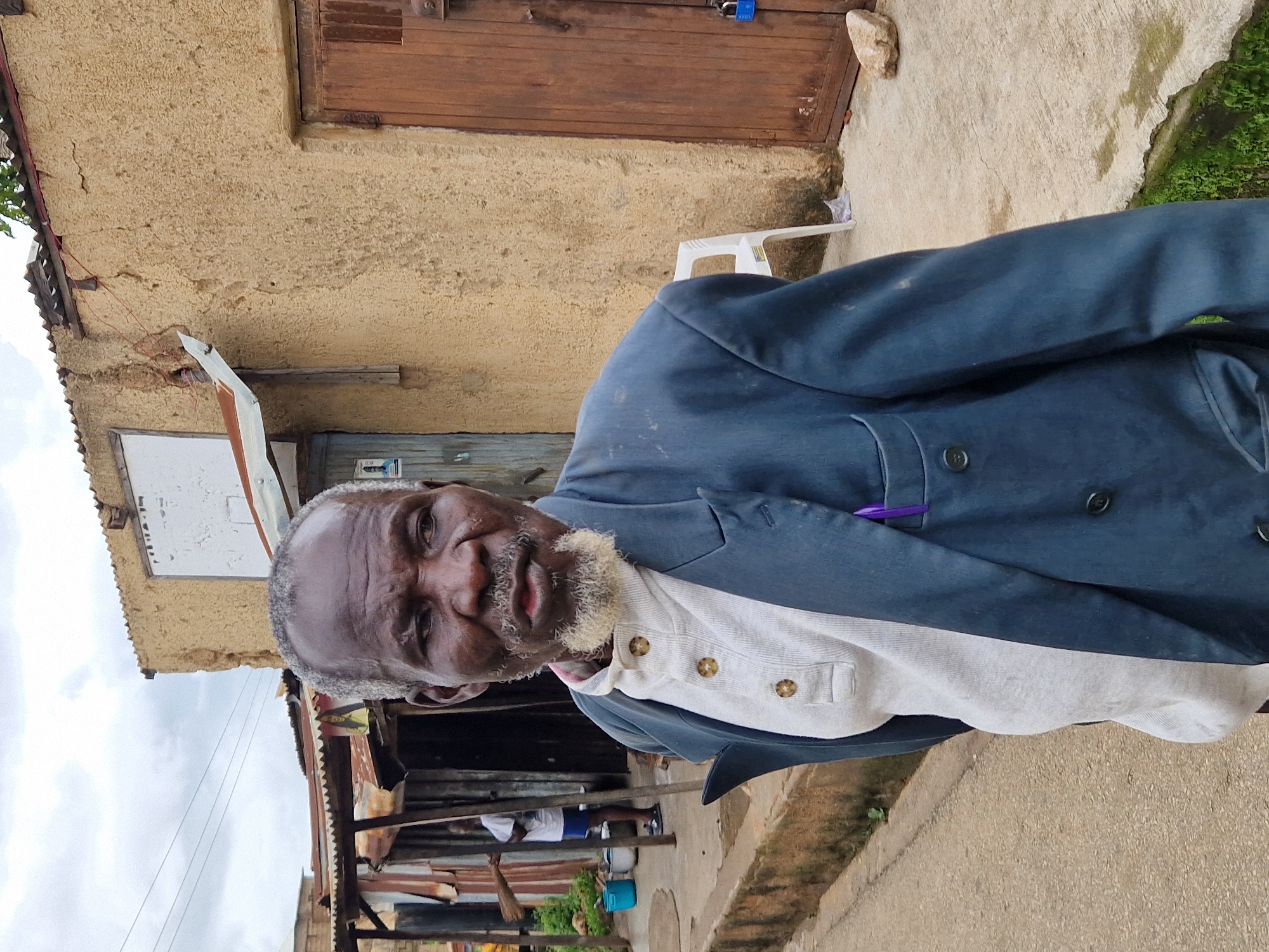 An outdoor, medium shot captures an elderly Black man standing on a concrete path in front of a building with a stucco facade in Jos, Nigeria. The man is facing slightly to his right and looking forward. He has short, gray hair, a gray beard, and appears to have dark skin. He is wearing a light gray collared shirt with three buttons and a dark blue double-breasted suit jacket. A purple pen is tucked into the breast pocket of his jacket. To the man's left, a corrugated metal awning extends from a building with rough, tan-colored stucco walls. A second, smaller building with a corrugated metal roof is visible behind the awning. To the man's right, a light-colored wooden door is set into the stucco wall. A blue padlock is attached to the door. A white plastic chair is positioned in front of the door. In the background, another person is visible under the corrugated metal awning, holding a broom. The ground is concrete with some visible cracks and patches of green moss. The sky is overcast.