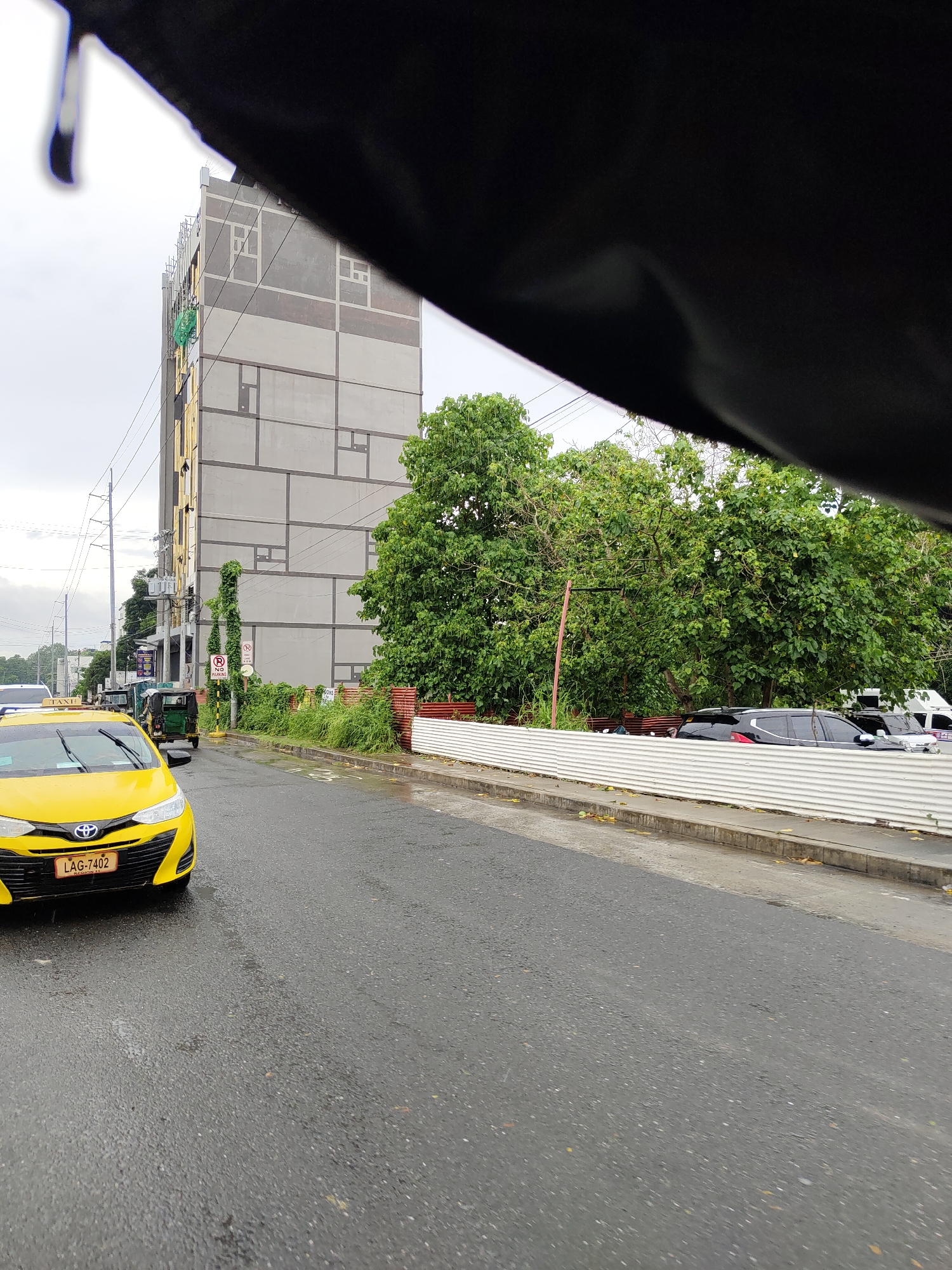 The image depicts a street scene in Davao City, Philippines, likely during the day, as indicated by the overcast sky. In the foreground, a yellow taxi with license plate "LAG-7402" is visible, its front partially cut off by the bottom of the frame. The road surface is wet, suggesting recent rain, with some scattered fallen leaves. A tall, modern building with a geometric facade is prominent in the background, under construction or recently completed, with scaffolding and green netting visible on its side. Adjacent to the building are lush green trees and vegetation. To the right, a corrugated metal fence separates the road from the properties behind it. Traffic is present, with another vehicle, possibly a tricycle, visible behind the taxi. The overall atmosphere is that of a typical urban street environment in the Philippines, with a mix of modern architecture and abundant greenery.