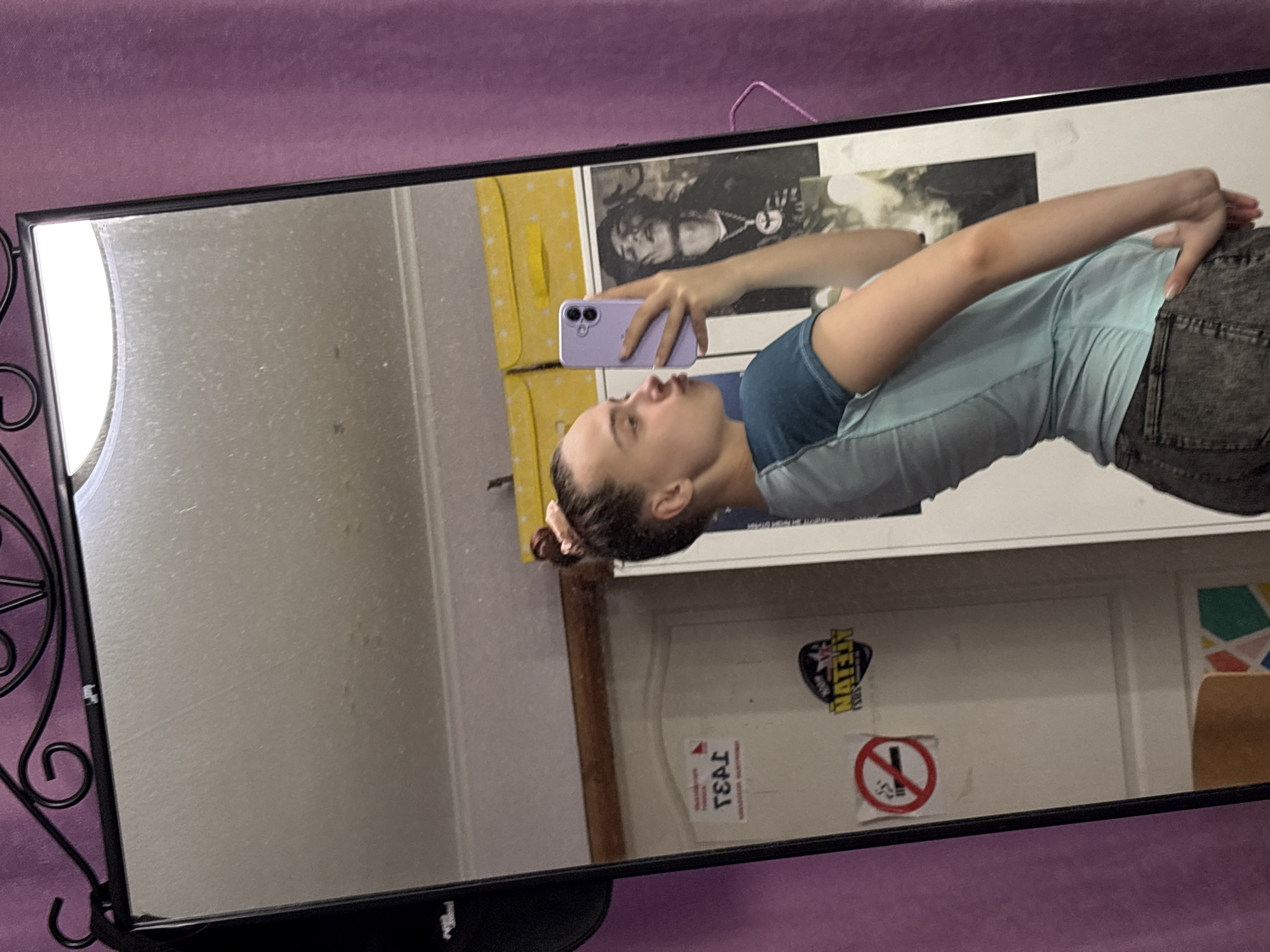 A person is taking a mirror selfie. The mirror is black with ornate details on the left side. The person is wearing a blue t-shirt and gray pants. The person is holding a purple cell phone. The background includes a poster with faces, a yellow storage container, and a white door with stickers. There is a pink sheet in the background. The location might be Прикамский, Russia.