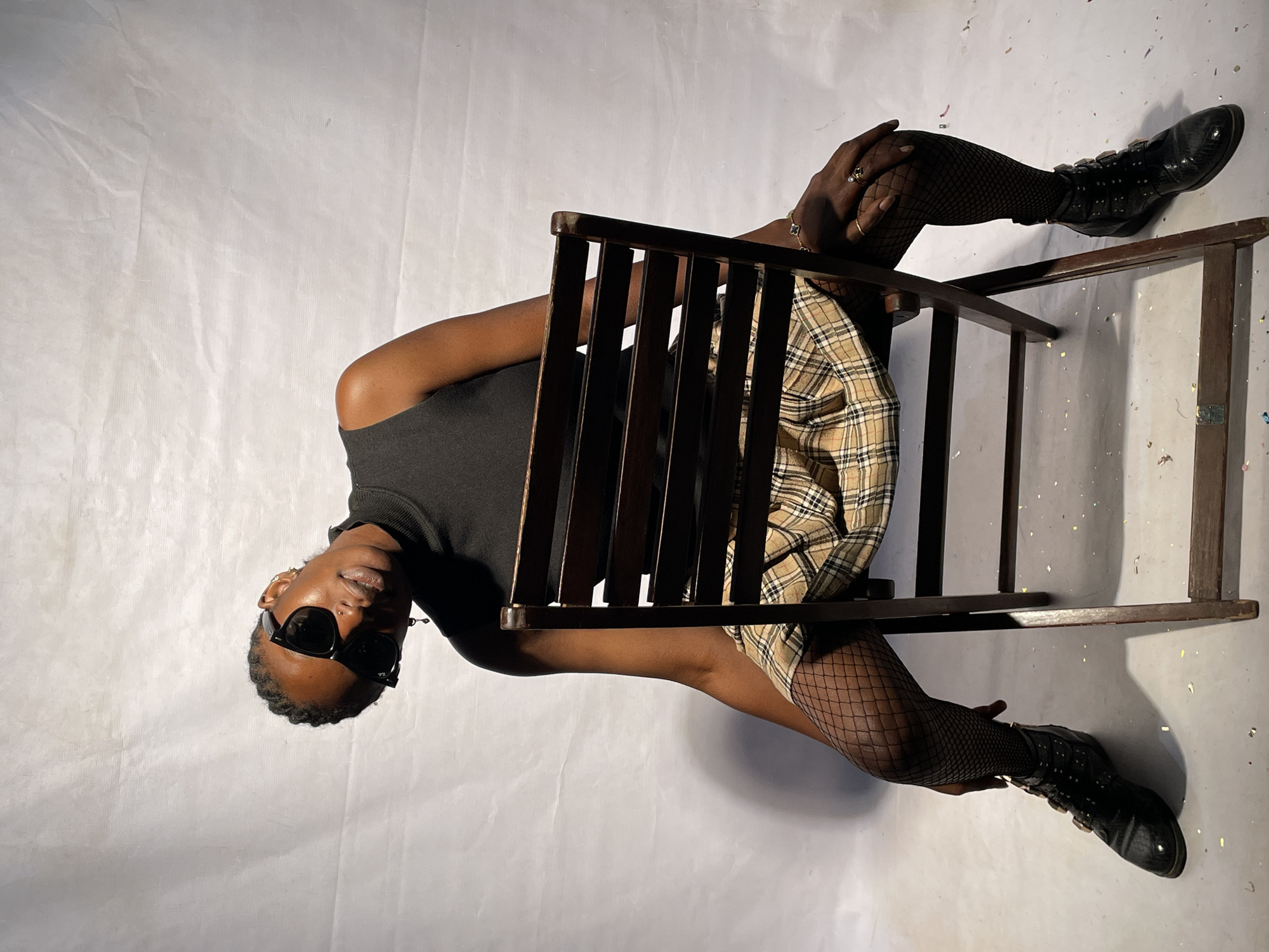 A person wearing a black turtleneck shirt, a tan plaid skirt, fishnet stockings, and black boots is seated in a wooden chair against a white background with confetti scattered on it. The person is also wearing black sunglasses and has jewelry on their hands. The person is tilted slightly to their right and is looking at the viewer. There is no indication of the city of Jos, Nigeria.