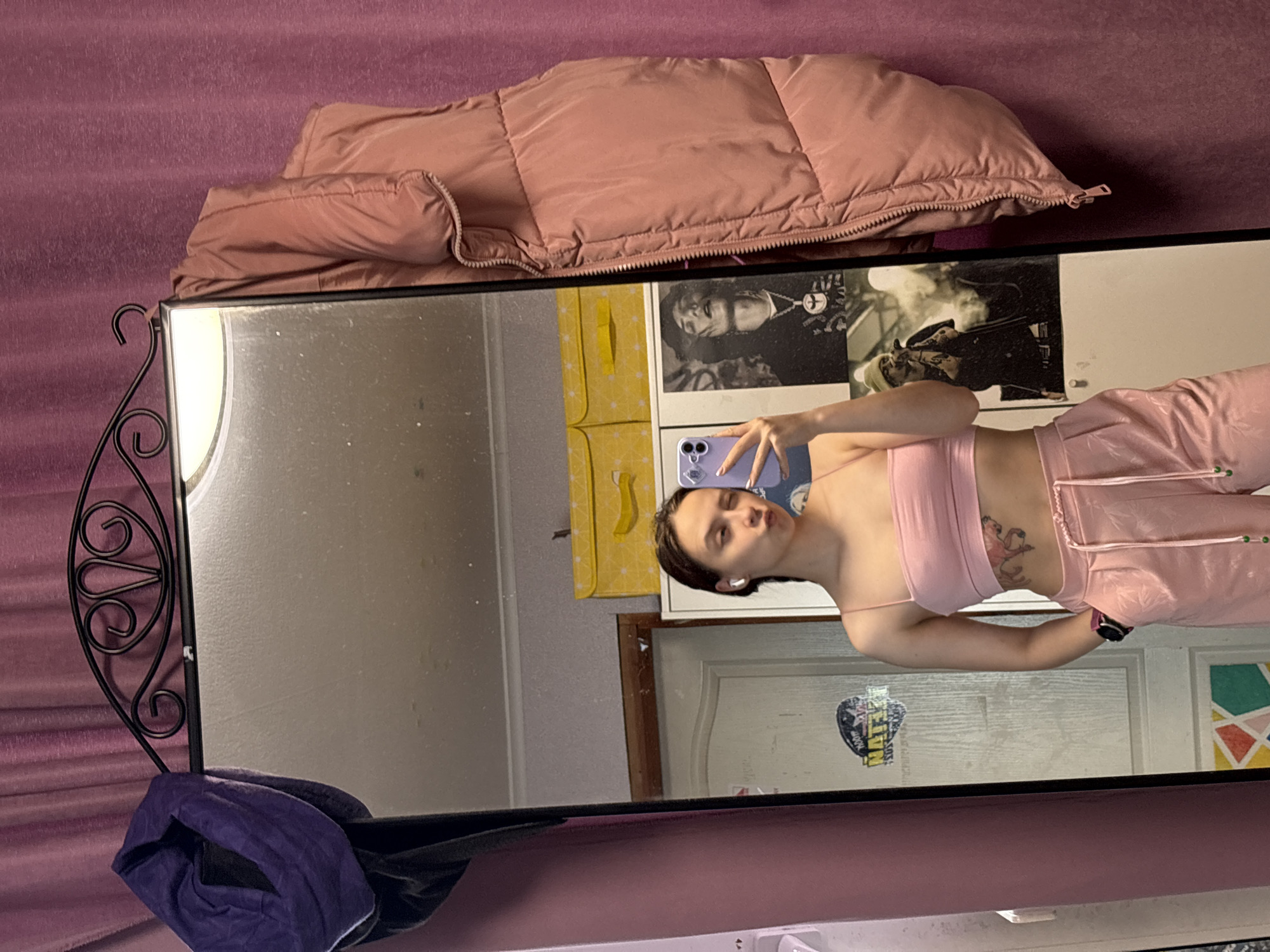 The image captures a young woman taking a mirror selfie indoors, likely in a private room. She is reflected in a large, rectangular mirror with a decorative black metal frame on the left. The woman has short, dark hair, is wearing a light pink crop top, matching shiny pink pants, and white wireless earbuds. A dark watch or bracelet is visible on her left wrist, and a tattoo of a pink cartoonish figure is on her right ribcage. She holds a light purple smartphone in her right hand to take the photograph, displaying a neutral expression.
The background behind the mirror is a muted purple fabric, possibly a curtain or wall. A pink puffer jacket is folded on a surface, likely a bed, above the mirror. Behind the woman's reflection, various personal items are visible on the wall, including a yellow fabric item, two black and white posters featuring people, and a partially visible white door with a sticker. A dark purple cloth is draped over the bottom left of the mirror frame. The setting suggests a personal residence in Прикамский, Russia. No specific time of day or weather is discernible. Text on the posters and a door sticker is present but largely illegible, though "RETURN" might be visible on the door sticker.