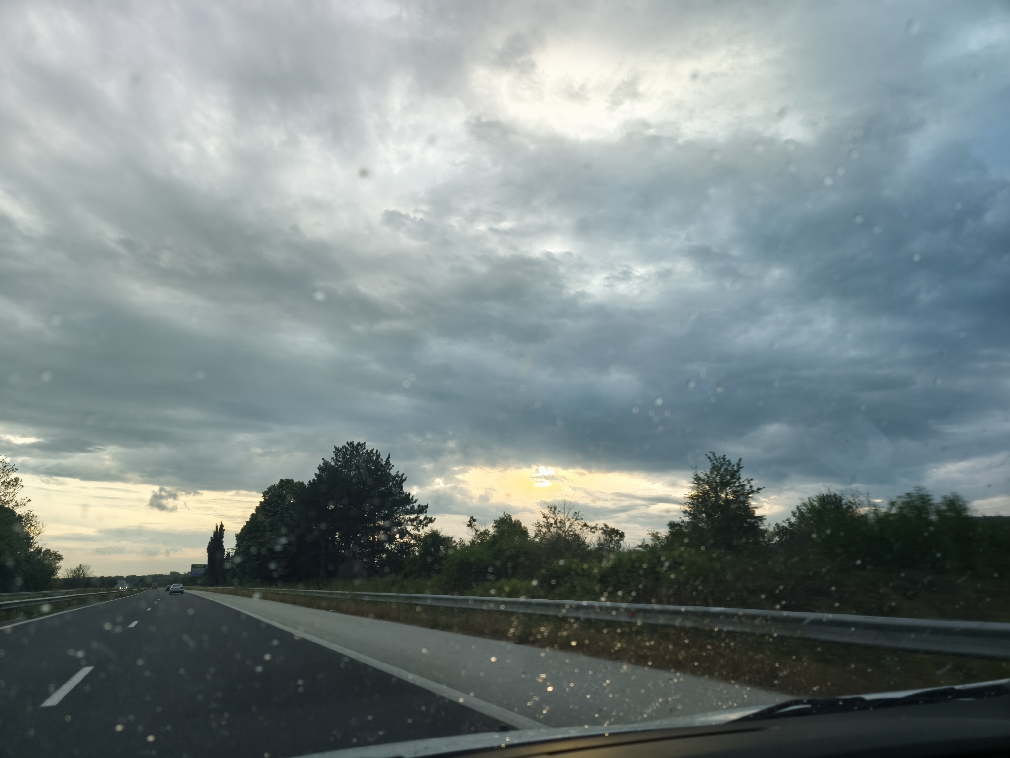 The image captures a first-person perspective from inside a moving vehicle on a multi-lane highway, looking forward through a windshield spattered with numerous small, blurry specks, possibly raindrops or dust. The sky dominates the upper portion of the frame, filled with dense, dramatic grey and bluish clouds, suggesting overcast weather or the approach of a storm. However, a bright, yellowish glow on the horizon, particularly evident on the right side and hinted at in the center, indicates the presence of a low sun, suggesting either late afternoon or early morning.
The highway, dark asphalt with white lane markings, stretches into the distance, where a few vehicles can be seen, including a white car farther ahead. A metallic guardrail runs along the right side of the road, separating it from a line of diverse trees and dense green foliage that extends along the horizon. On the far left, more distant trees are visible against the lighter sky. No specific location cues or discernible text are visible. The overall atmosphere conveys a sense of travel under a moody sky with hints of breaking light.