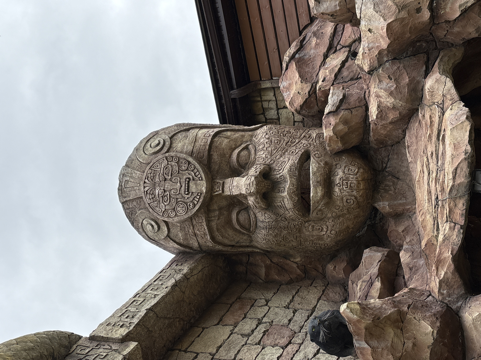 The image shows a large stone sculpture of a human face, possibly Mayan or Aztec-inspired, located in Gelendzhik, Russia. The face is intricately carved with detailed patterns and a circular headpiece. The sculpture is attached to a building facade featuring stone-like textured panels and some dark wooden elements near the top. The sky is overcast and visible in the background. A decorative rock formation is present on the right side of the face, and a black, round, object that resembles a spotlight is present at the bottom of the image.