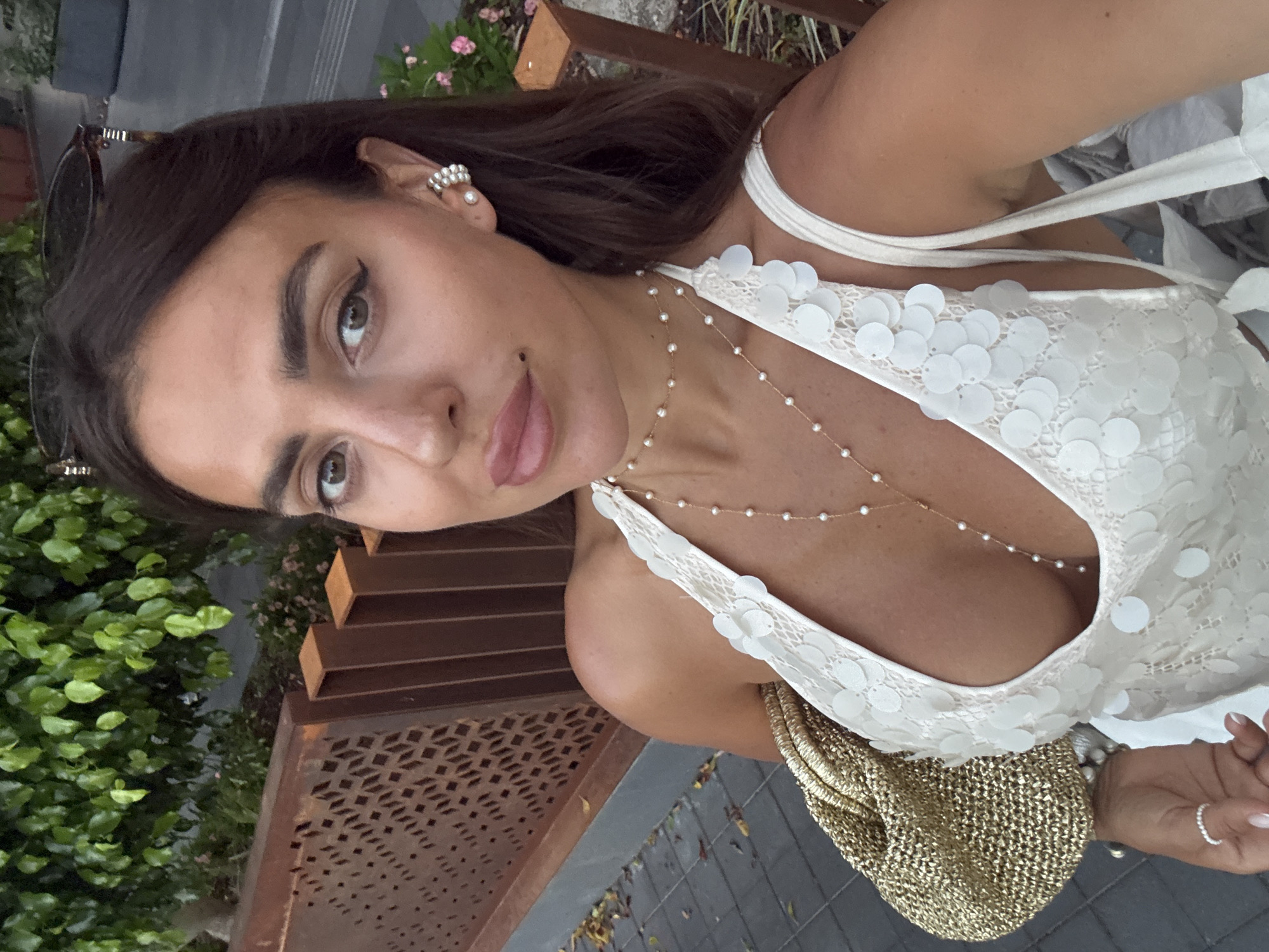 A woman is captured in a close-up, selfie-style photograph, with the camera held at an angle that emphasizes her décolletage and upper body. She is wearing a white dress adorned with circular white sequins and lace detailing, with thin straps. A delicate pearl necklace is draped across her chest. In her left hand, she holds a textured gold bag and wears a thin silver ring on her ring finger. Her right ear is visible, featuring a prominent pearl earring.
The background suggests an outdoor setting. To the left of the frame, lush green foliage is visible, hinting at a garden or natural environment. To the right, there are elements of an urban or built environment, including a dark, textured surface that appears to be paving stones and a reddish-brown structure with an ornate, perforated pattern. The image is shot from a low angle, looking upwards towards the woman's face. Her eyes are directed towards the camera, and she has a slight smile.
The lighting suggests it is daytime, possibly late afternoon or early evening, with a warm, natural light illuminating the scene. The overall mood is relaxed and fashionable, with the woman appearing confident and poised. The image is taken in Fethiye, Turkey, as indicated by the location context.