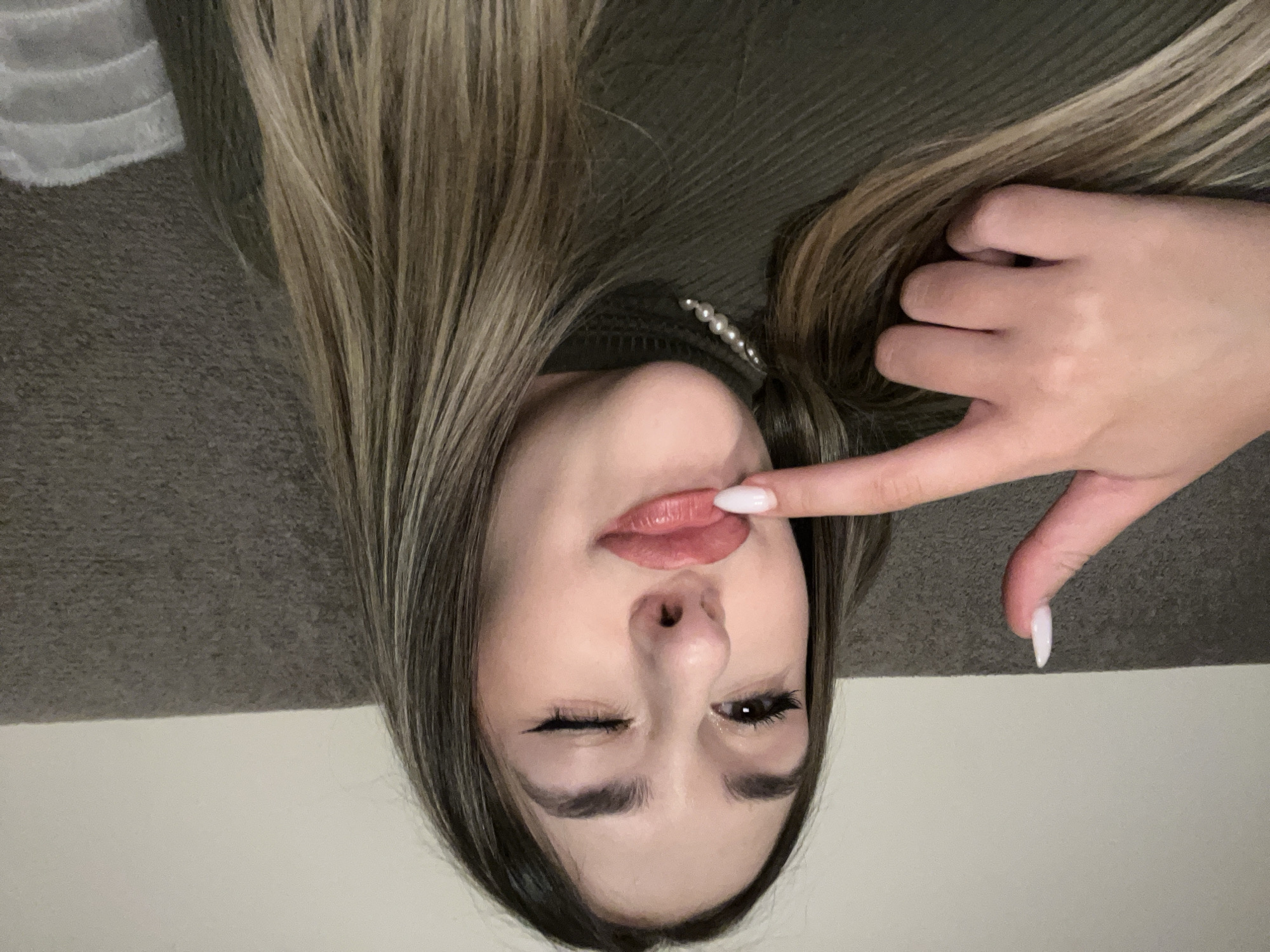 A close-up, rotated portrait of a young woman taken indoors, likely in Sofia, Bulgaria. The woman is positioned with her head to the left and her body extending to the right of the frame, giving the impression she is lying down. She has long, straight hair with visible highlights, possibly light brown or blonde streaks, cascading across a dark green ribbed top or sweater. A string of pearls is visible near her neck.
Her face is at the center of the image, featuring a playful expression. Her left eye is closed in a wink, while her right eye is open. She has dark eyeliner with a winged shape and mascara on her eyelashes. Her lips are slightly parted and appear to have lip gloss or lipstick. Her left index finger, with a long, pointed white nail, is touching her lower lip. Her skin tone is fair.
The background consists of a dark gray, textured surface, possibly a blanket or upholstery, in the upper portion of the image, and a lighter, plain surface, possibly a wall or floor, in the lower portion. The lighting suggests an indoor setting, and no specific time of day can be determined. There is no visible text.