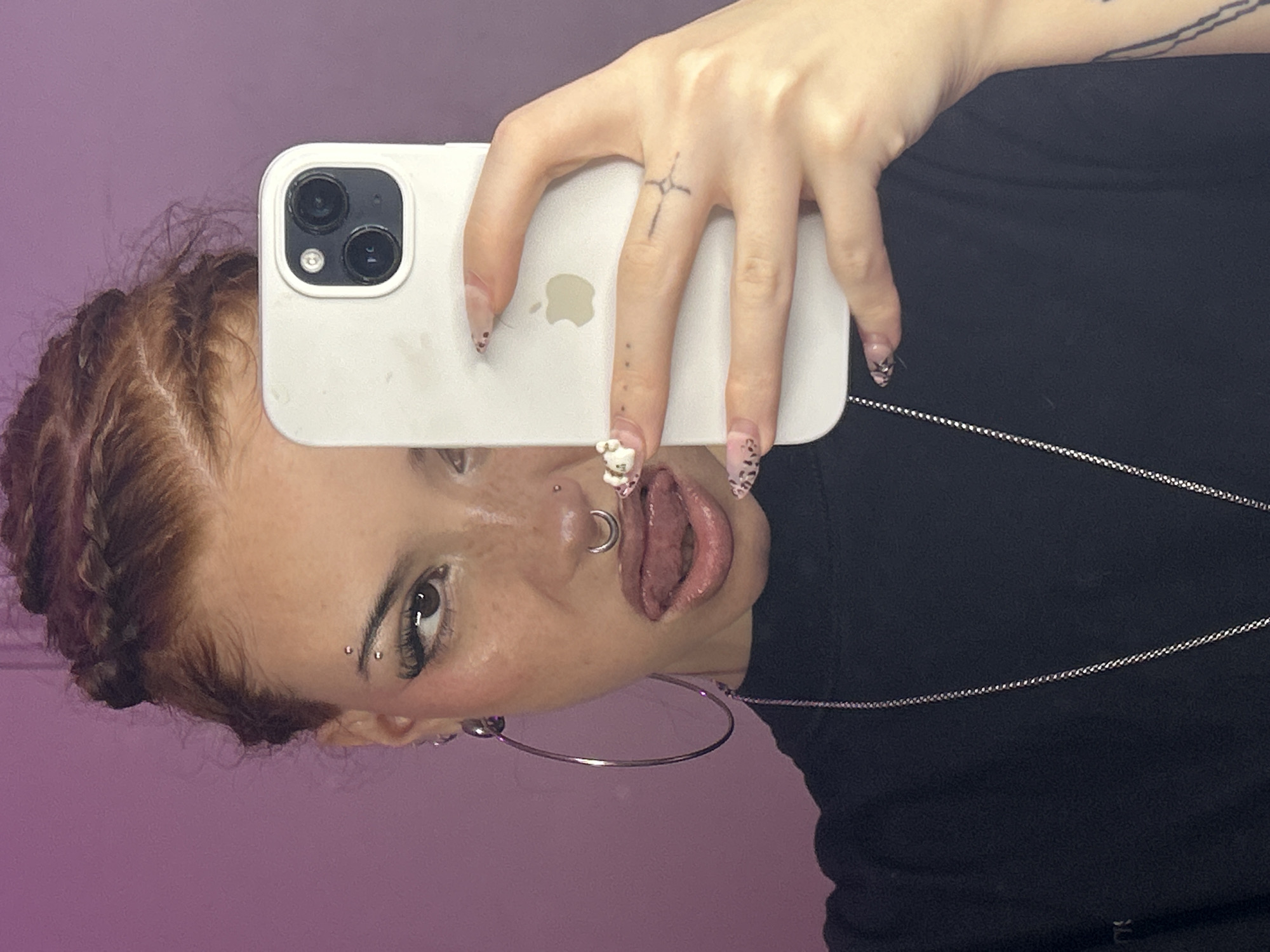 A young woman takes a selfie in front of a lavender background. She holds a white iPhone in her right hand. Her hair is reddish-brown and braided. She has piercings above her eyebrow and in her nostril. She sticks out her tongue. She wears a black shirt and a silver chain necklace. There is no indication of the location as Buenos Aires, Argentina.