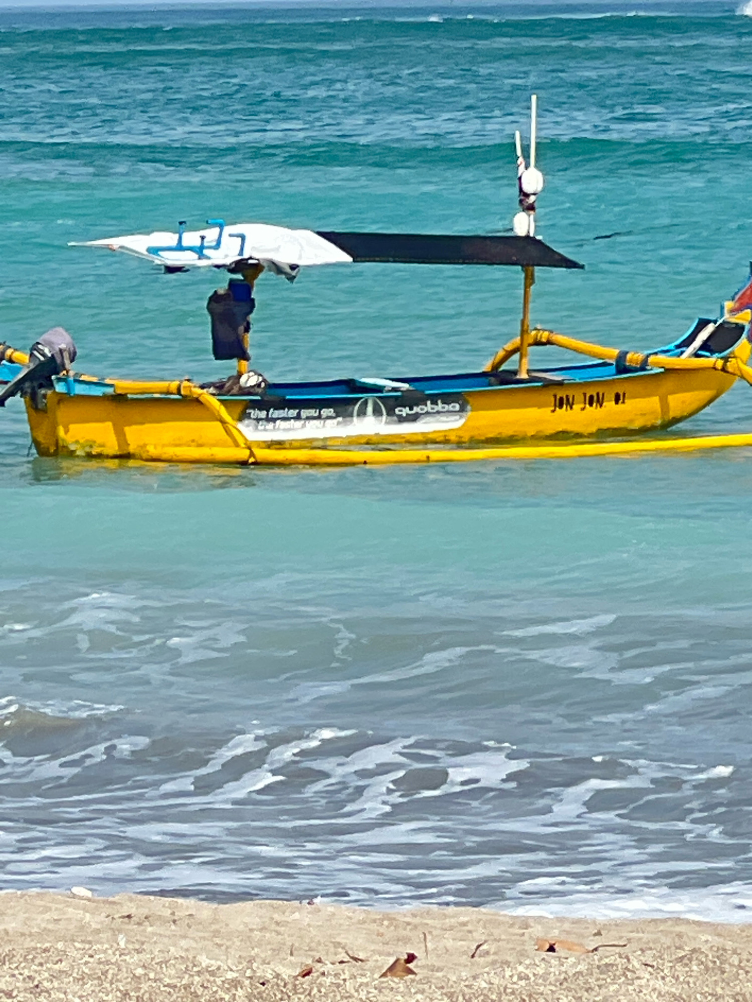 In Kuta, Indonesia, a yellow outrigger canoe named "JON JON 01" floats on turquoise waters near a sandy beach. The boat has a blue stripe along its gunwale and a white awning with blue trim supported by a black canopy structure. On the left side of the boat, a black outboard motor is attached, and a dark bag is tied to the stern. The boat also features a white sticker with the word "quobba" and text that reads "the faster you go, the faster you go". The ocean is calm with gentle waves, and the sky is clear. The scene captures a peaceful moment at the popular Kuta beach.