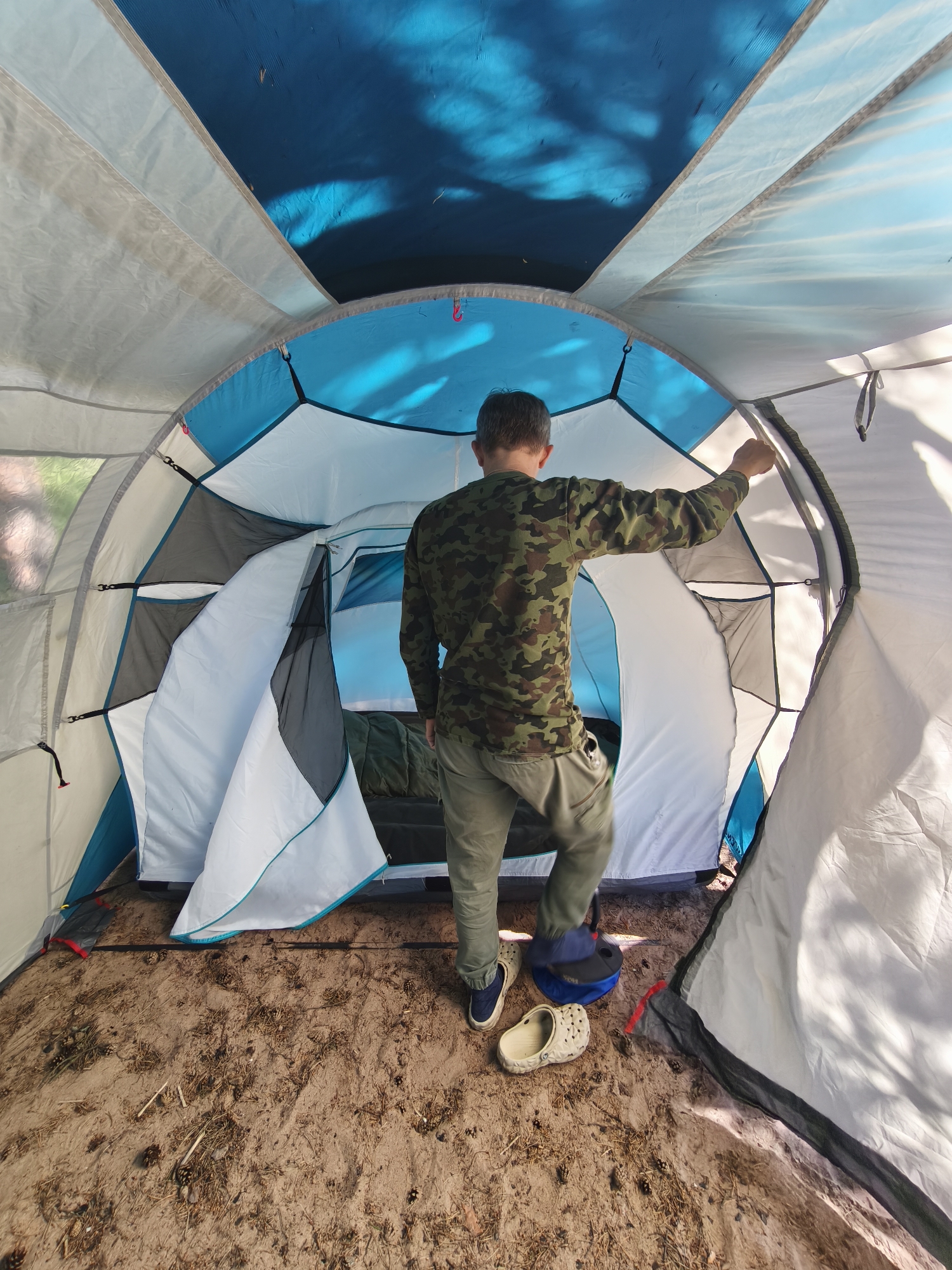 A boy in a camouflage shirt and khaki pants stands inside a large tent, his back to the camera. He is holding open the tent flap with his right hand, looking towards the interior. The tent is spacious, with white, blue, and gray fabric. The floor is covered in sand with scattered pinecones and twigs. Inside the tent, a sleeping bag is visible on what appears to be a cot. Two pairs of shoes, one pair of blue sneakers and a pair of light-colored Crocs, are on the ground near the entrance. The sun is shining, casting dappled shadows on the tent's interior, suggesting it is daytime. The location appears to be a campsite, given the tent and natural ground cover. The overall mood is one of outdoor activity, likely camping or preparing to camp. There is no visible text in the image.
