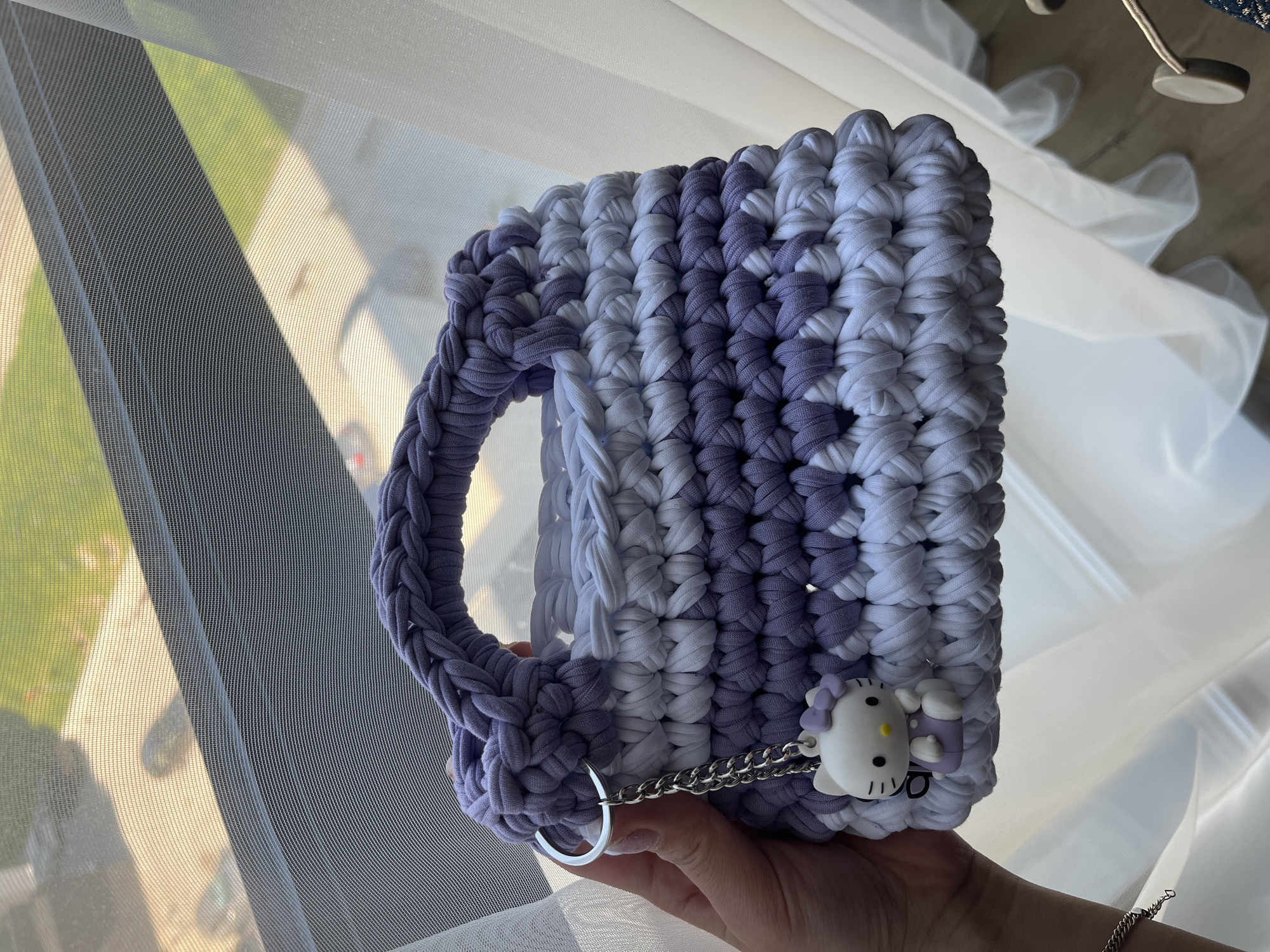 A hand holds a crocheted purse in front of a window. The purse is made of thick yarn in alternating stripes of lavender and white. It has a rounded handle on the left side and a small Hello Kitty charm hanging from a silver chain on the lower right corner. The hand holding the purse is pale.
Through the window, a blurry view of a neighborhood is visible. There's greenery and a light-colored building. The scene is brightly lit by sunlight.
Due to the limited information within the image, the specific location within Plovdiv, Bulgaria, cannot be determined.
