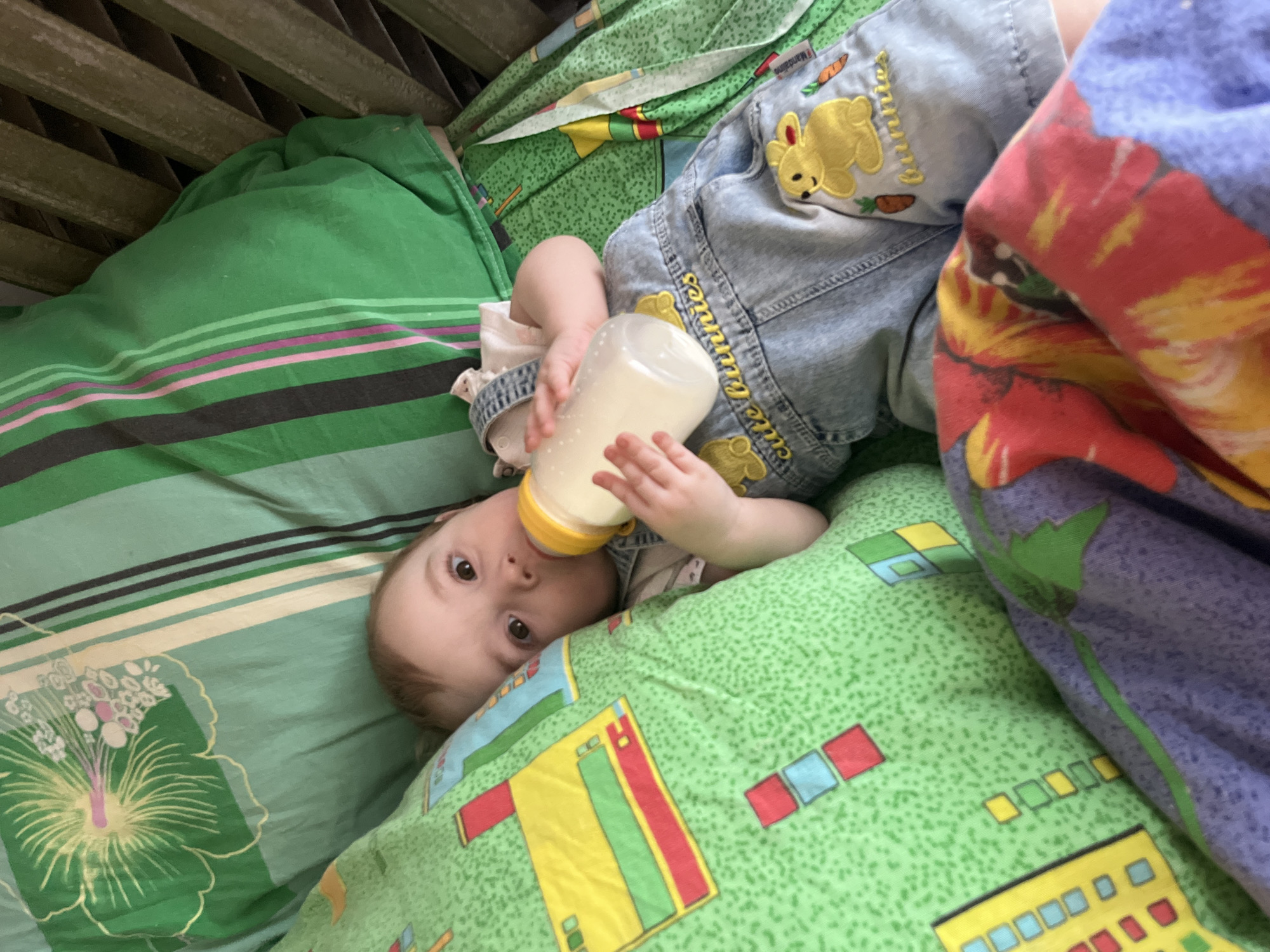 An infant is observed lying supine in a wooden crib within a residence in Романята, Russia. The infant, dressed in light-wash denim overalls featuring yellow bunny embroidered patches and "cute bunnies" text, holds a clear baby bottle with a yellow cap and nipple containing a white liquid. The crib is furnished with multiple textile items: a green pillow or blanket with a pattern of yellow, red, and blue geometric shapes on the left, a green sheet with a large yellow and white floral design and vertical green, black, and pink stripes on the top and upper right, and a folded multicolored blanket with purple, red, and yellow abstract patterns in the lower right foreground. Wooden crib slats are visible on the right side of the image. The infant's eyes are open and directed towards the viewer.