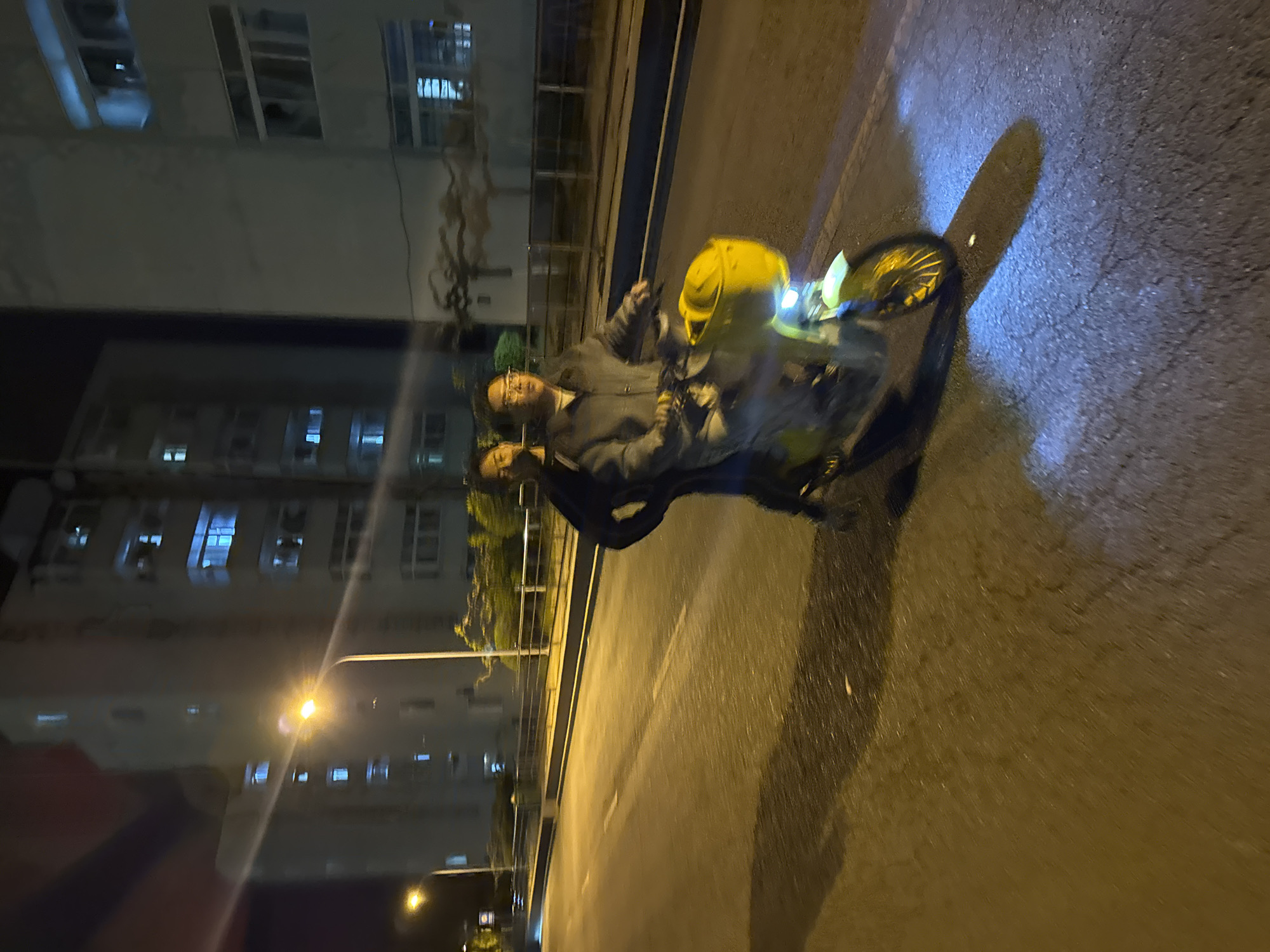 Two people are on a motorcycle at night. The location appears to be in the city of Zhongtun, China, based on the building style. The buildings are lit up at night. The road is wet.