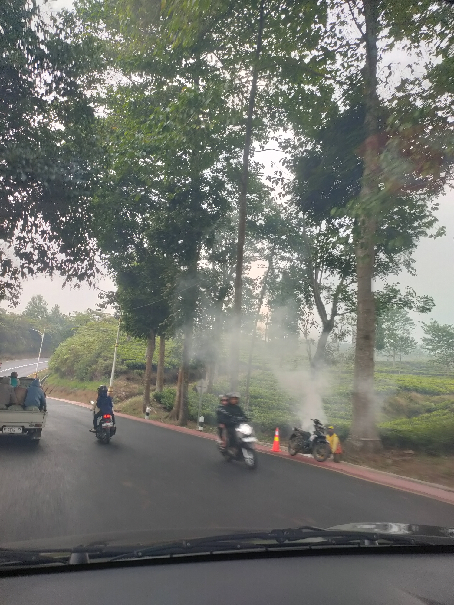 The image depicts a roadside scene in what is indicated to be the vicinity of Subang, Indonesia, likely a rural or semi-rural area with a prominent tea plantation. The perspective is from inside a vehicle, looking forward through the windshield. A paved road runs through the scene, with a white truck carrying sacks of goods on the left side, traveling in the same direction as the viewer. On the road, there are two motorcycles. The first, closer to the truck, is ridden by a person wearing a dark blue jacket and helmet. Further ahead, a second motorcycle, white in color, is ridden by two people, one of whom appears to be wearing a dark helmet.
To the right of the road, there is a distinct orange traffic cone. Behind it, a dark-colored motorcycle is parked, and a person in a yellow jacket is standing next to it, with what appears to be smoke or steam rising from the ground or the motorcycle. Beyond the road and to the right, there is a vast expanse of green tea plantation, characteristic of the region. Tall trees line the left side of the road, with dense foliage and a light sky visible through them. The overall atmosphere is overcast or hazy.