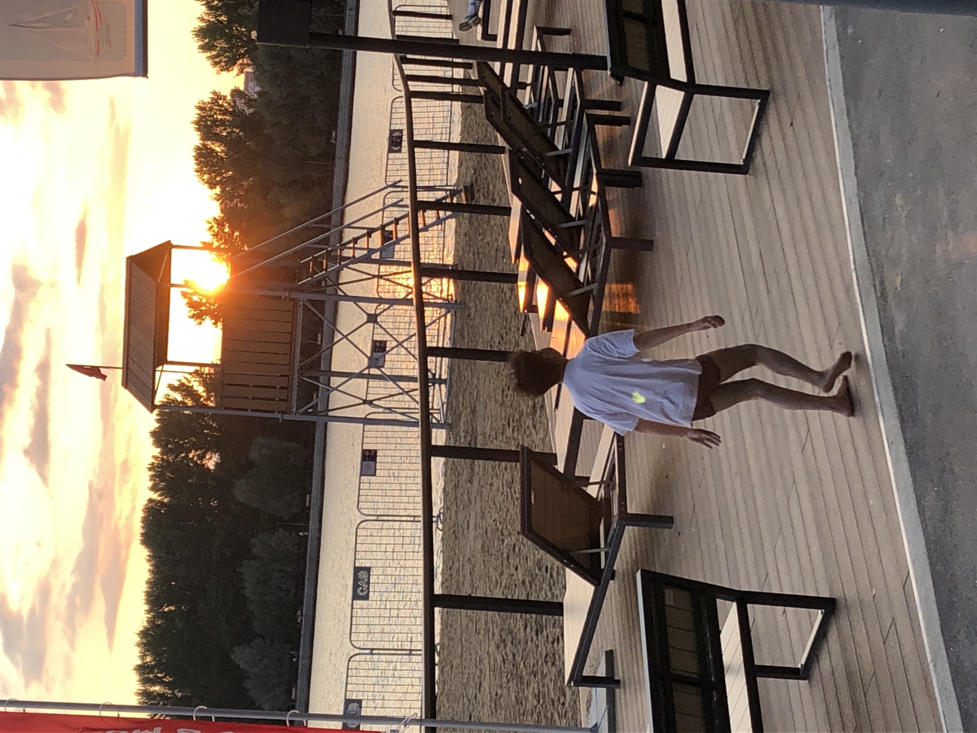 The image depicts a scene at a waterfront leisure area in Khimki, Russia, during sunset. A young person, viewed from behind, is walking on a wooden deck towards the right side of the frame. They are wearing a white t-shirt and brown shorts. The sun is setting behind a lifeguard tower with stairs, casting a warm glow and highlighting the water. Several empty lounge chairs are arranged on a sandy beach area in the mid-ground, separated from the deck by a metal fence. Trees line the horizon behind the beach. The overall atmosphere is tranquil, with the fading light creating long shadows.