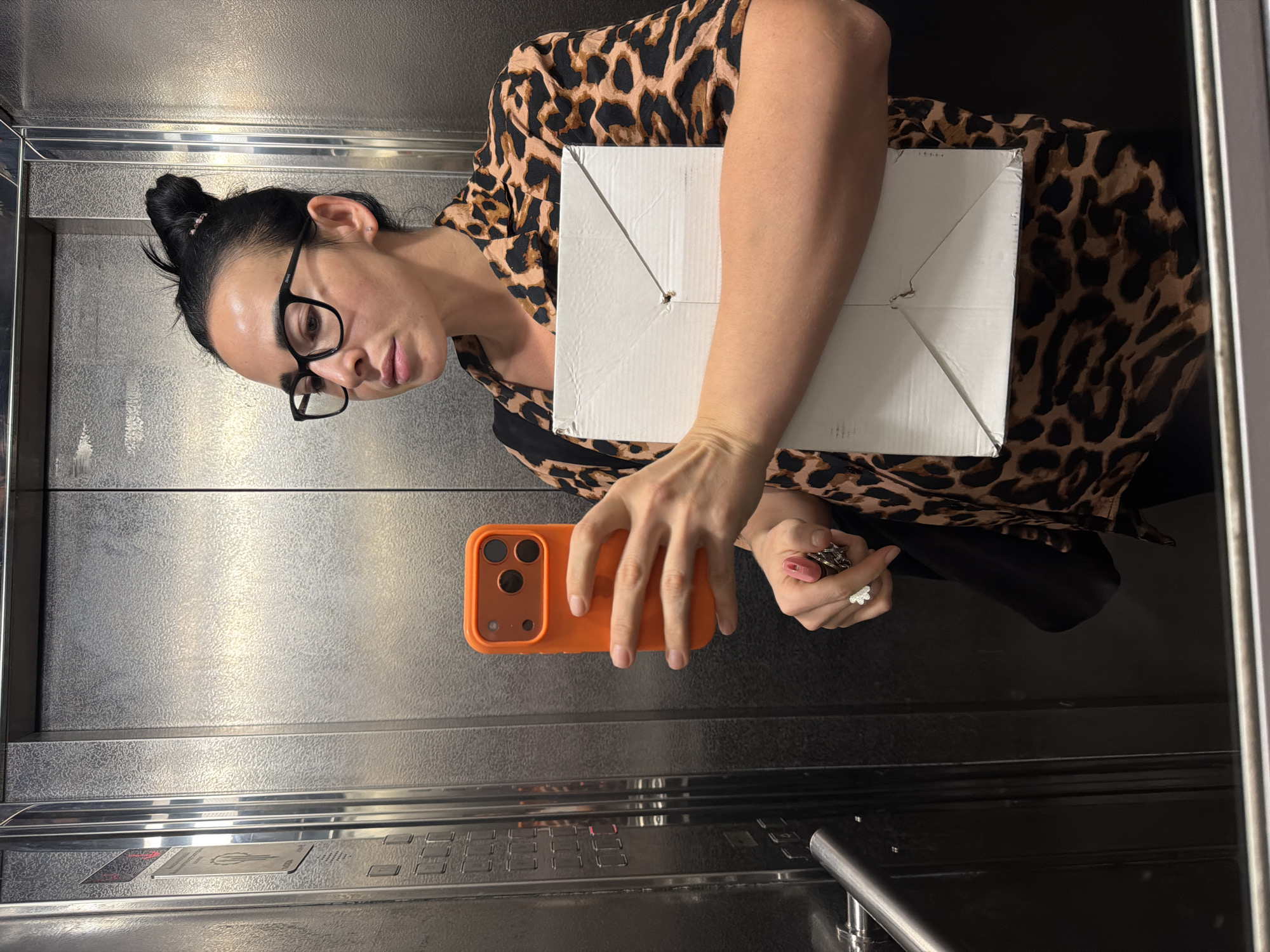 An adult female is depicted taking a mirror selfie inside an elevator in the city of Unknown, Russia. She has dark hair styled in a messy bun, wears black-framed glasses, and a brown and black leopard-print collared shirt. In her right hand, she holds an orange-cased smartphone with a prominent three-lens camera array, positioned to capture the reflection. Her left hand holds a flat, rectangular white cardboard box against her midsection and a partially open tube of pink lipstick. A silver-toned ring shaped like a flower is visible on her left ring finger. The elevator interior features textured metallic panels on the walls. To the left in the reflection, an elevator control panel is visible with dark square buttons and red indicator lights. A metallic handrail is present at the bottom left of the image.