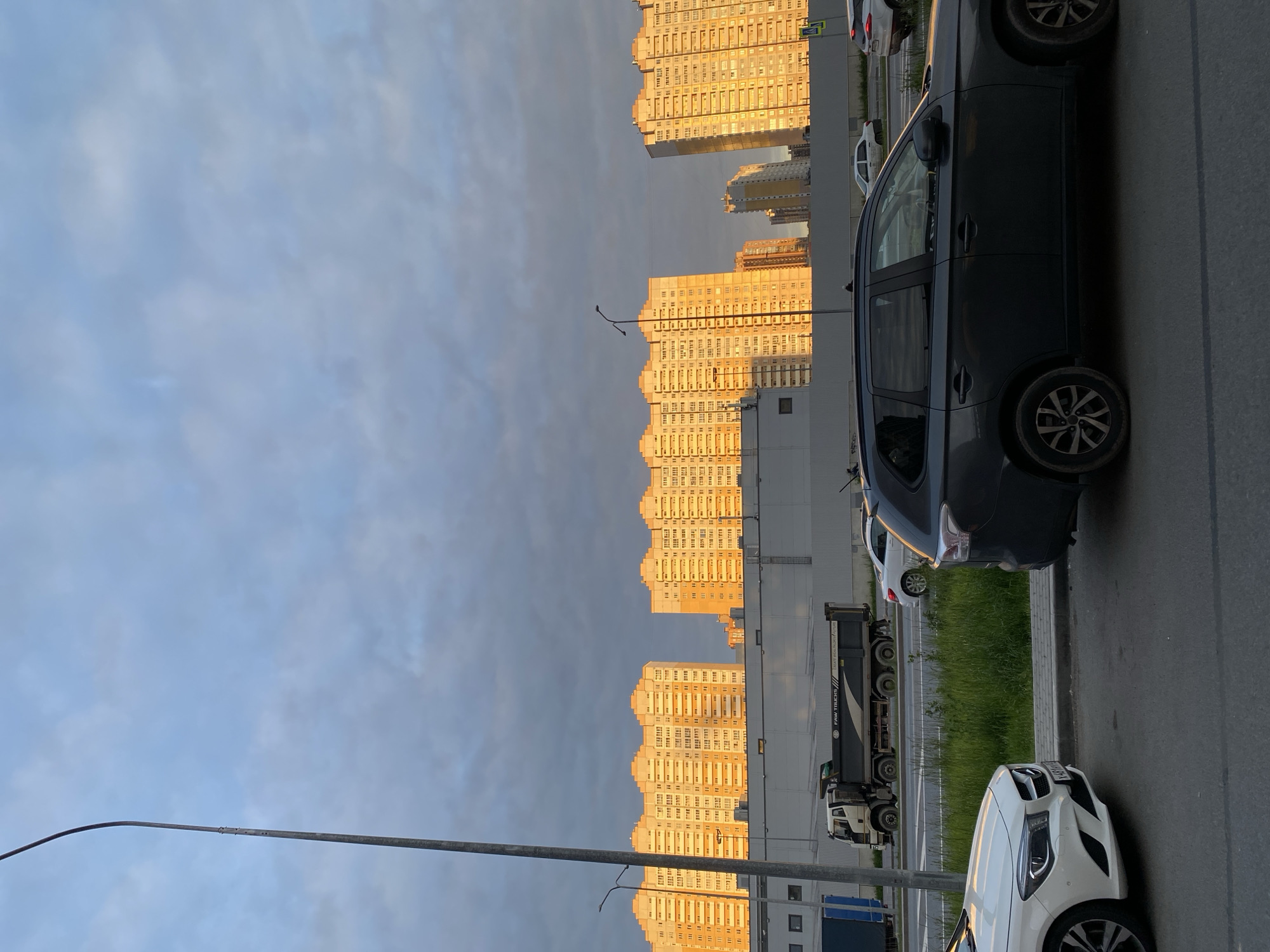 This image depicts an urban street scene during what appears to be the golden hour, with warm, low-angle sunlight illuminating the background.
The primary subjects are large, multi-story residential buildings that line the far side of a wide street. These buildings, characterized by their repeating patterns of windows and balconies, are bathed in a strong, golden light, causing them to appear yellow or orange despite the overall cloudy sky. Their scale suggests a high-density residential area.
In the foreground and mid-ground, a paved road and sidewalk run parallel to the buildings. Several vehicles are present. A dark-colored SUV, possibly a Mitsubishi Outlander, is prominently featured on the right side of the road. Further back, a white Mercedes-Benz sedan is visible with its front and driver's side showing, bearing the license plate "A464KH 799". A large black dump truck, with "FAW TRUCKS" printed on its side, is also visible. Other cars are partially seen, indicating a busy street. A strip of green grass separates the road from the buildings in some sections.
The sky, occupying a significant portion of the upper left frame, is overcast with soft, greyish-blue clouds, suggesting partial cloud cover that still allows the sun to break through. A tall, slender street lamp post with a curved arm stands on the left side of the frame. The time of day is notable for the strong directional lighting, creating dramatic contrast between the sun-drenched buildings and the shadowed road and foreground elements. There is no specific activity or event taking place beyond the normal flow of traffic and parked cars in a city environment.