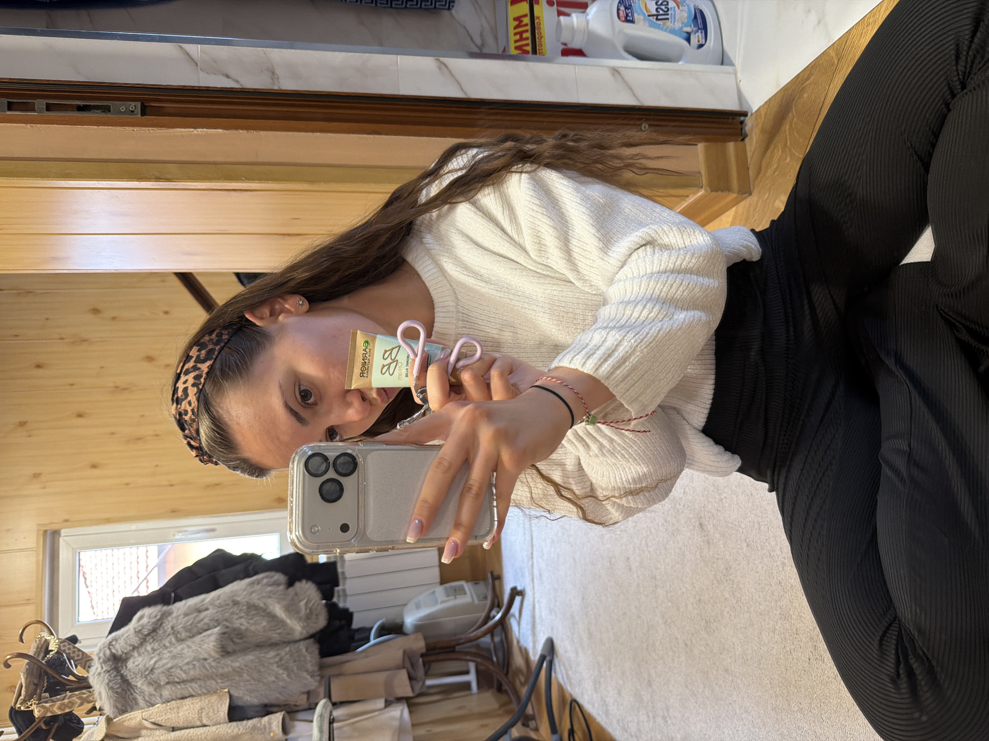 A young woman with long brown hair and a leopard-print headband is taking a selfie. She is wearing a white sweater and black leggings. She is holding a silver iPhone with a clear case and a green and yellow tube of Garnier BB cream. She is holding a pair of pink hair clips with her hand. The background is a room with wood paneling and a window. A rack of clothes is reflected in a mirror behind her. A bottle of laundry detergent is visible in the top corner. The location is indicated as Buhovo, Bulgaria.