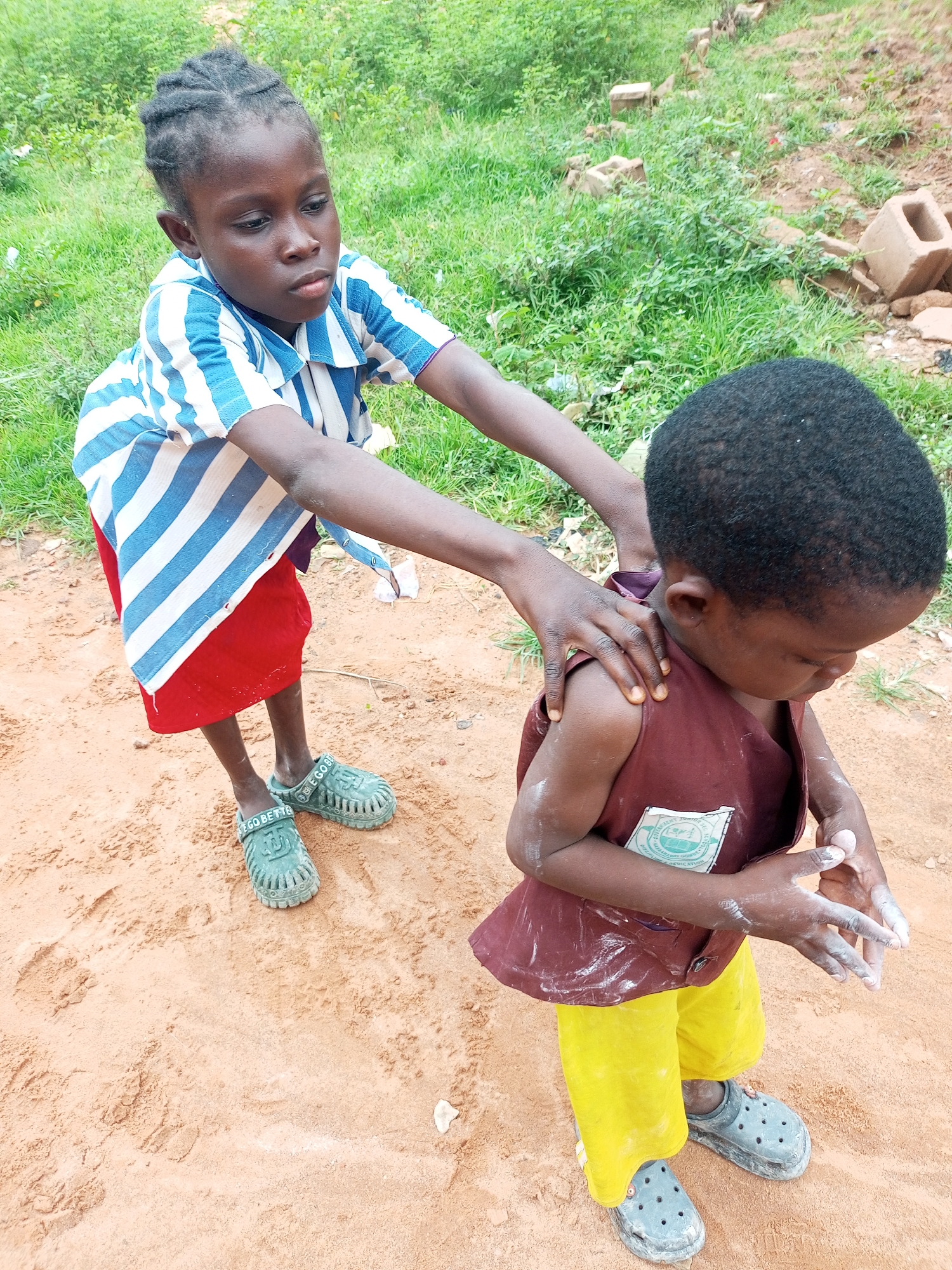 Two children, one older and one younger, are standing on sandy ground in what appears to be a rural or semi-rural area. The older child, on the left, is wearing a blue and white striped shirt and a red skirt. They are reaching out and touching the shoulders of the younger child. The younger child, on the right, is wearing a maroon sleeveless vest over yellow shorts. Their hands are clasped together in front of them, and they appear to be looking down at their hands. The background consists of green vegetation and some scattered bricks and debris. The overall impression is one of everyday life in an outdoor setting.