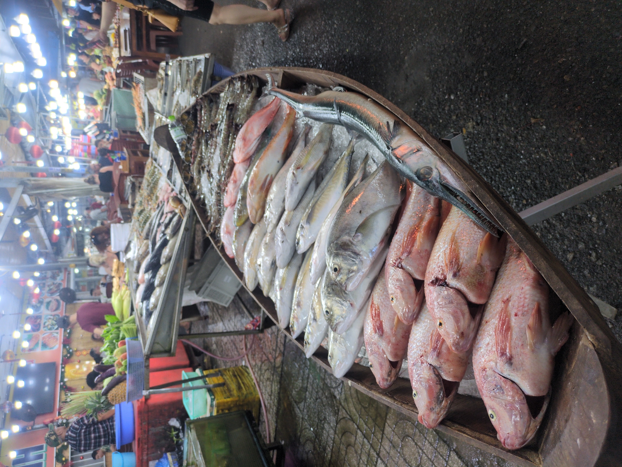 The image depicts a fish market in Phú Quốc, Vietnam. Various types of fish, including red tilapia, are displayed on ice in a wooden boat-shaped container for sale. Behind the fish display, there are other food vendors and customers. The market is illuminated by overhead lights. Vegetables, fruits, and other seafood are visible in the background. The ground appears to be wet. The overall scene is a busy outdoor market.