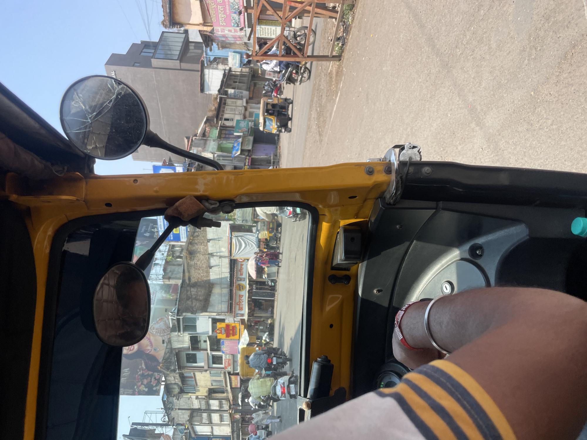 The image provides a frontal view from inside a yellow auto-rickshaw on a commercial street in Kolhapur, India, under clear daylight conditions.
In the immediate foreground, the right arm and shoulder of an individual, likely the auto-rickshaw driver, are visible on the left. The person wears a light-colored shirt with dark horizontal stripes. A silver bangle and a red and white patterned bracelet are present on the wrist. Below the arm, a segment of the vehicle's black dashboard and steering column is observed, with a key inserted into the ignition. A teal-colored bottle cap is partially visible at the bottom edge. The yellow frame of the auto-rickshaw occupies a significant portion of the left and top-right sections, framing the street view. Two rearview mirrors extend from the frame: the left mirror is intact and reflects a billboard advertisement, while the right mirror exhibits extensive starburst cracking across its surface.
The midground reveals an asphalt road with visible wear. Vehicular traffic includes multiple motorcycles and scooters, with a white scooter directly ahead and a person on a motorcycle to the right. Another yellow auto-rickshaw is present further down the road, moving away from the viewer. Pedestrians are visible on the sidewalks alongside the road. A brown wooden barrier or fence structure is positioned on the right side of the road.
The background consists of a dense urban streetscape lined with multi-story buildings of varying ages and architectural styles, some appearing weathered. Numerous commercial establishments feature awnings and signage in both Marathi and English. Specific visible signs include "ADITYA," "MAHARAJAJ," and "फोलपुरी ड्रायफ्रुट्स वडा" (Kolhapuri Dryfruits Wada). A large billboard on the left side of the street prominently displays a smiling woman adorned with jewelry. Overhead power lines and utility poles are visible against a clear, bright blue sky. The scene depicts a bustling commercial thoroughfare.
