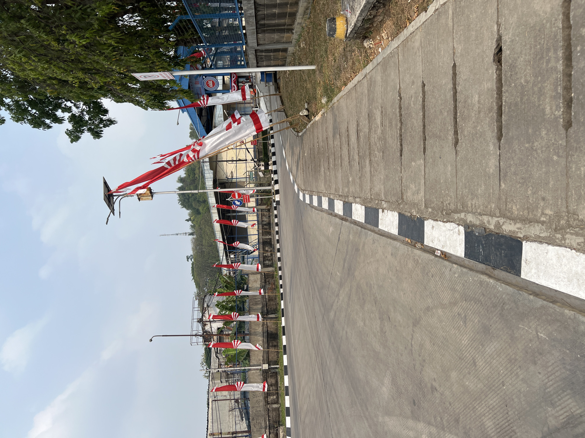 The image depicts a paved road with a sidewalk in Klapanunggal, Indonesia. The road has textured markings and is bordered by a curb painted with black and white stripes. Along the sidewalk, several Indonesian flags, a red and white motif, are planted in the ground, some appearing slightly tattered. The sidewalk itself is made of concrete slabs with visible drainage openings. In the background, there are industrial-looking buildings, a tall radio tower, and lush green trees. The sky is overcast. There are no people visible in the frame.