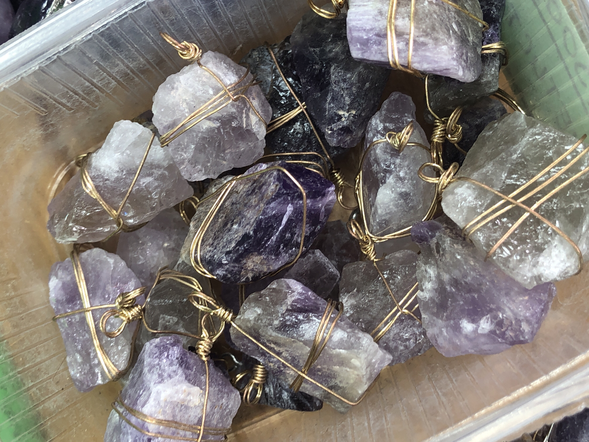 A close-up, high-angle shot shows a collection of raw, uncut amethyst crystals wrapped with gold-colored wire. The crystals vary in size and shade of purple, from pale lavender to deep violet, with some showing translucent areas. The wire is intricately wrapped around each stone, forming small loops and twists, suggesting they are intended for jewelry or decorative purposes. The crystals are piled together in a transparent plastic container, with the container's ribbed texture visible in the background. The lighting is bright, highlighting the facets and inclusions within the crystals. The scene is devoid of any people or specific environmental cues that would indicate its location in the city of Туим, Russia.
