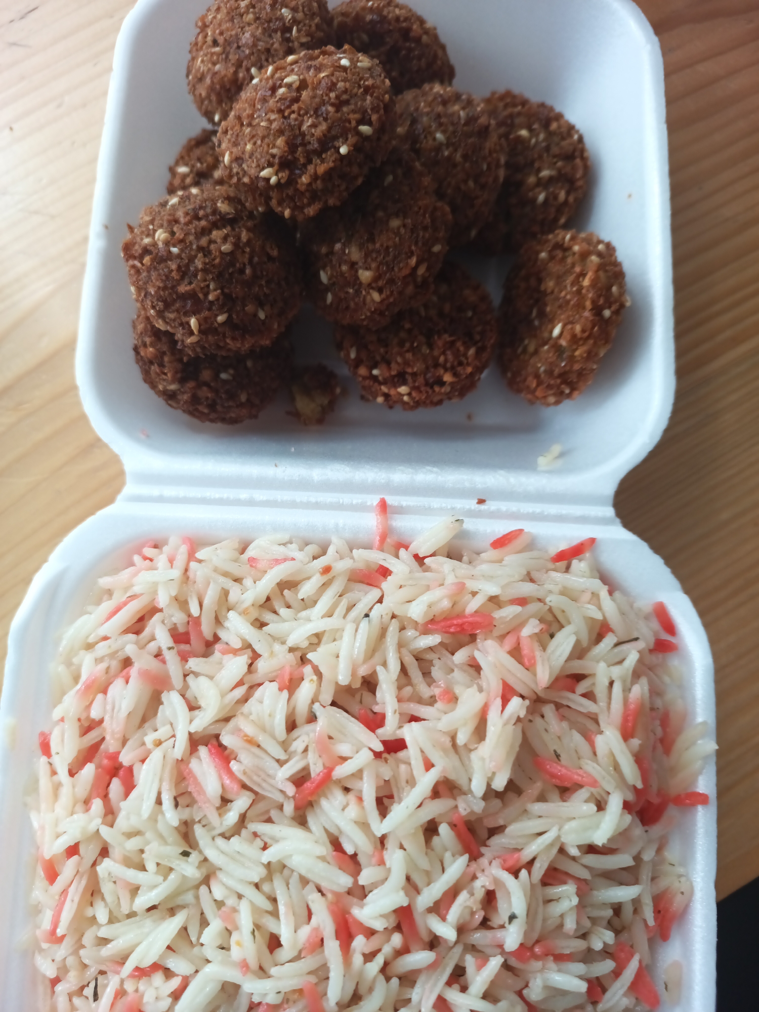 The image shows a white styrofoam takeout container divided into two sections. The top section contains a pile of approximately ten brown, spherical falafel, each speckled with sesame seeds. The bottom section is filled with white rice interspersed with pieces of red-colored rice. The container rests on a light-colored wooden surface. There is no additional information to suggest that this image was taken in the city of Burgas, Bulgaria. No people, actions, or interactions are visible in the frame.