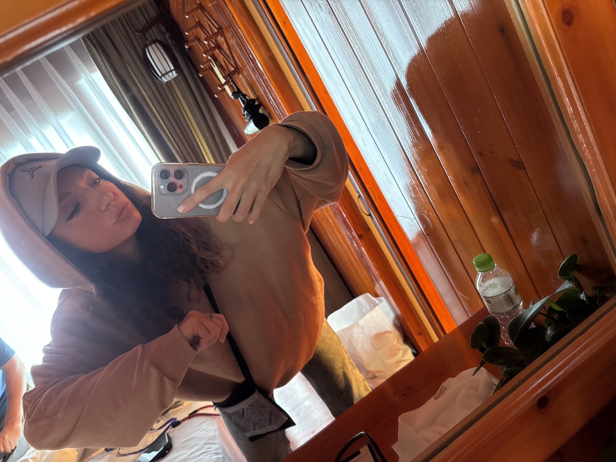 A woman in a tan hooded sweatshirt and a baseball cap is taking a selfie in a mirror. The reflection shows part of a bedroom with a bed, and a window with curtains. Reflected in the mirror's foreground is a water bottle and a small potted plant. The location appears to be in a hotel or room, and the background may suggest a location in Binh Lu, Vietnam, due to the style of the wood paneling and the decorative light fixture.