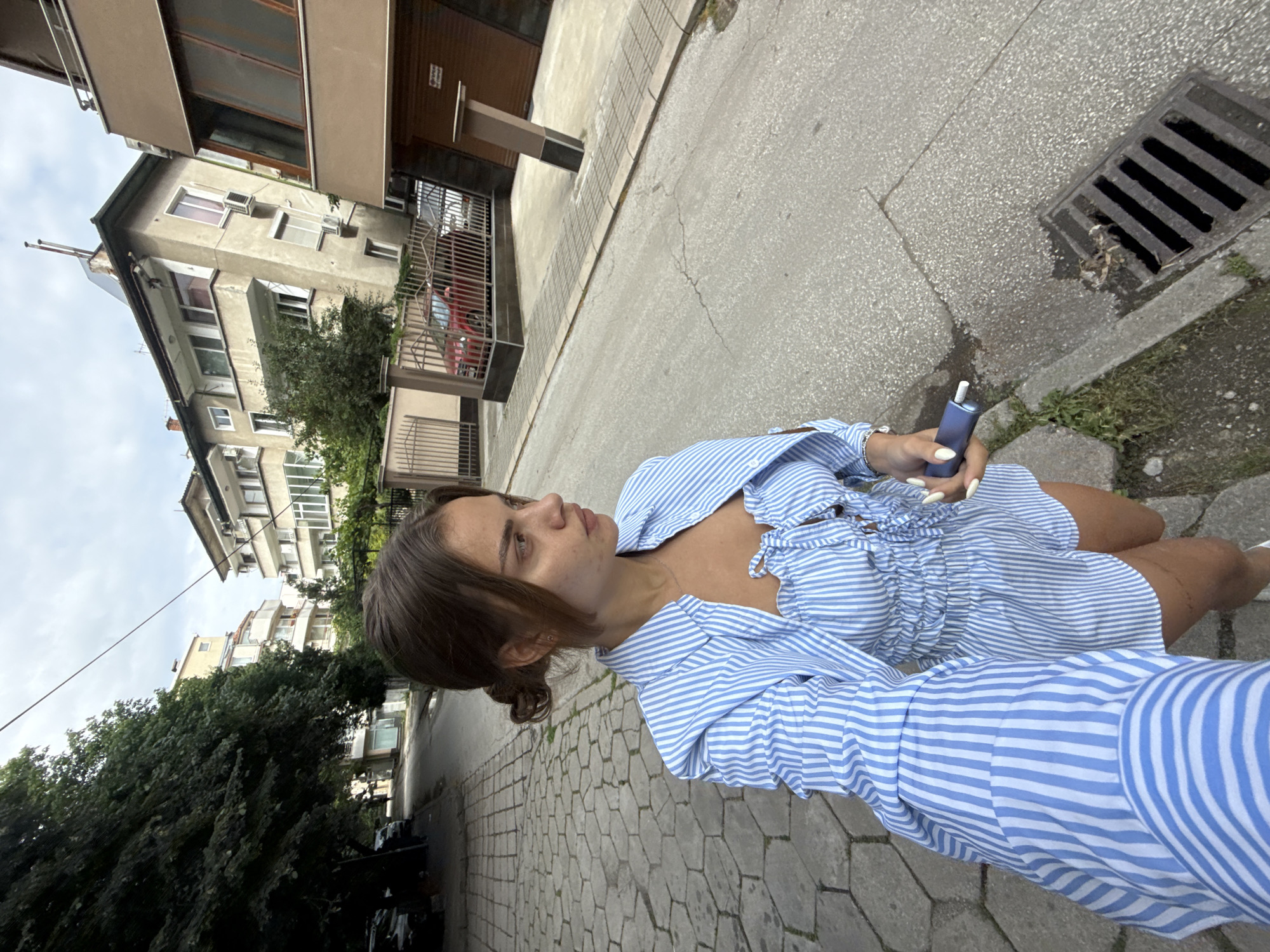 The image shows a young woman with brown hair pulled back, wearing a blue and white striped shirt. She is holding a blue electronic device in her right hand. The woman is outdoors, standing on a sidewalk paved with hexagonal stones. To her right, there is a street with a metal drain cover. In the background, there are buildings and trees. The buildings appear to be residential, with multiple stories and windows. There is also a red car visible behind a metal gate. The sky is overcast, suggesting it may be a cloudy day. The overall scene appears to be an urban street, possibly in a residential neighborhood. The woman's expression is neutral, and she is looking off to the side. The image is oriented sideways, with the top of the frame on the left. There is no visible text in the image.