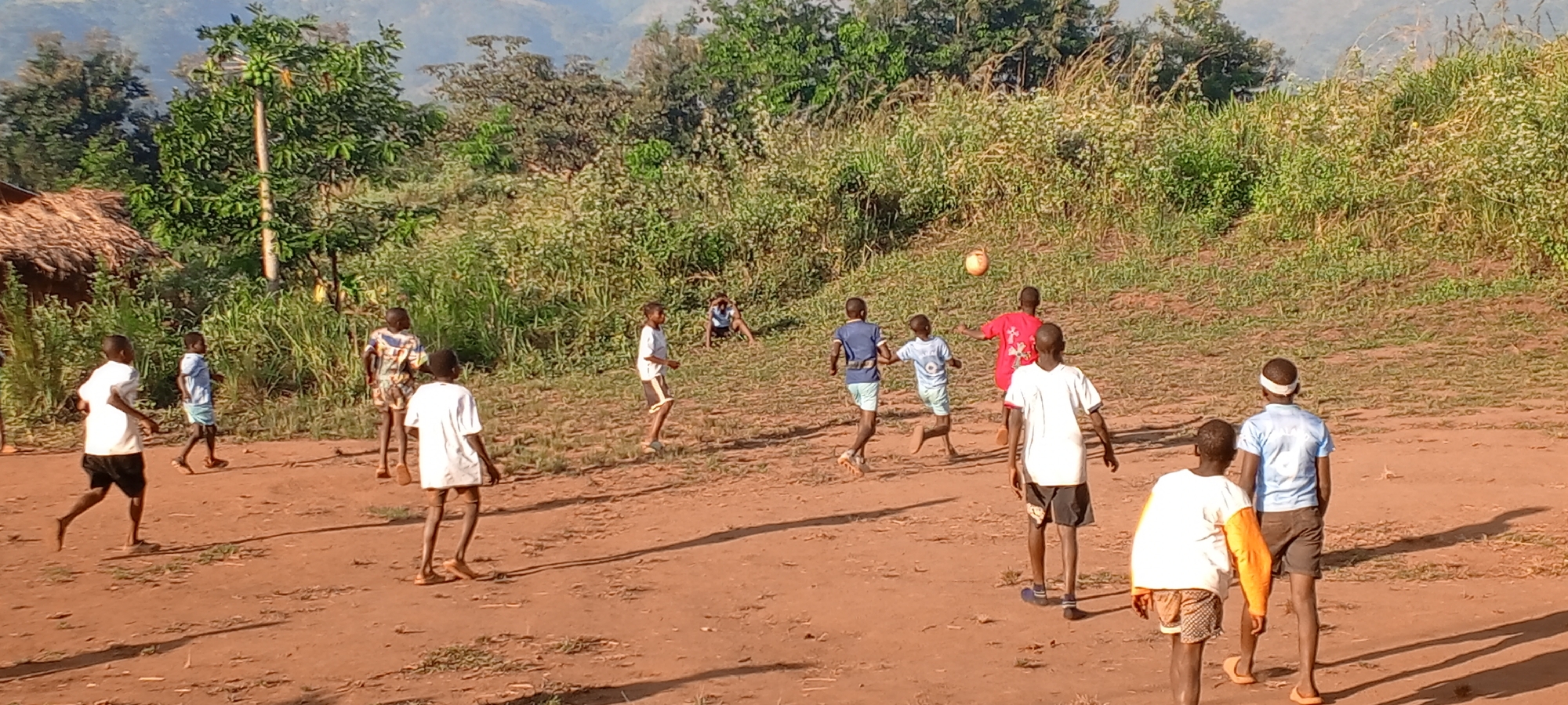 The image depicts a group of children playing soccer on a reddish-brown dirt field. The location is possibly near the city of Ngalula, Democratic Republic of the Congo, given the context. The field is situated in front of a backdrop of dense green vegetation and rolling hills. The children are wearing a variety of casual clothing, including t-shirts and shorts, some with bare feet. The soccer ball is airborne in the center of the group, with some children running towards it. There is a small building with a thatched roof visible on the left side of the image. The lighting suggests it is daytime.