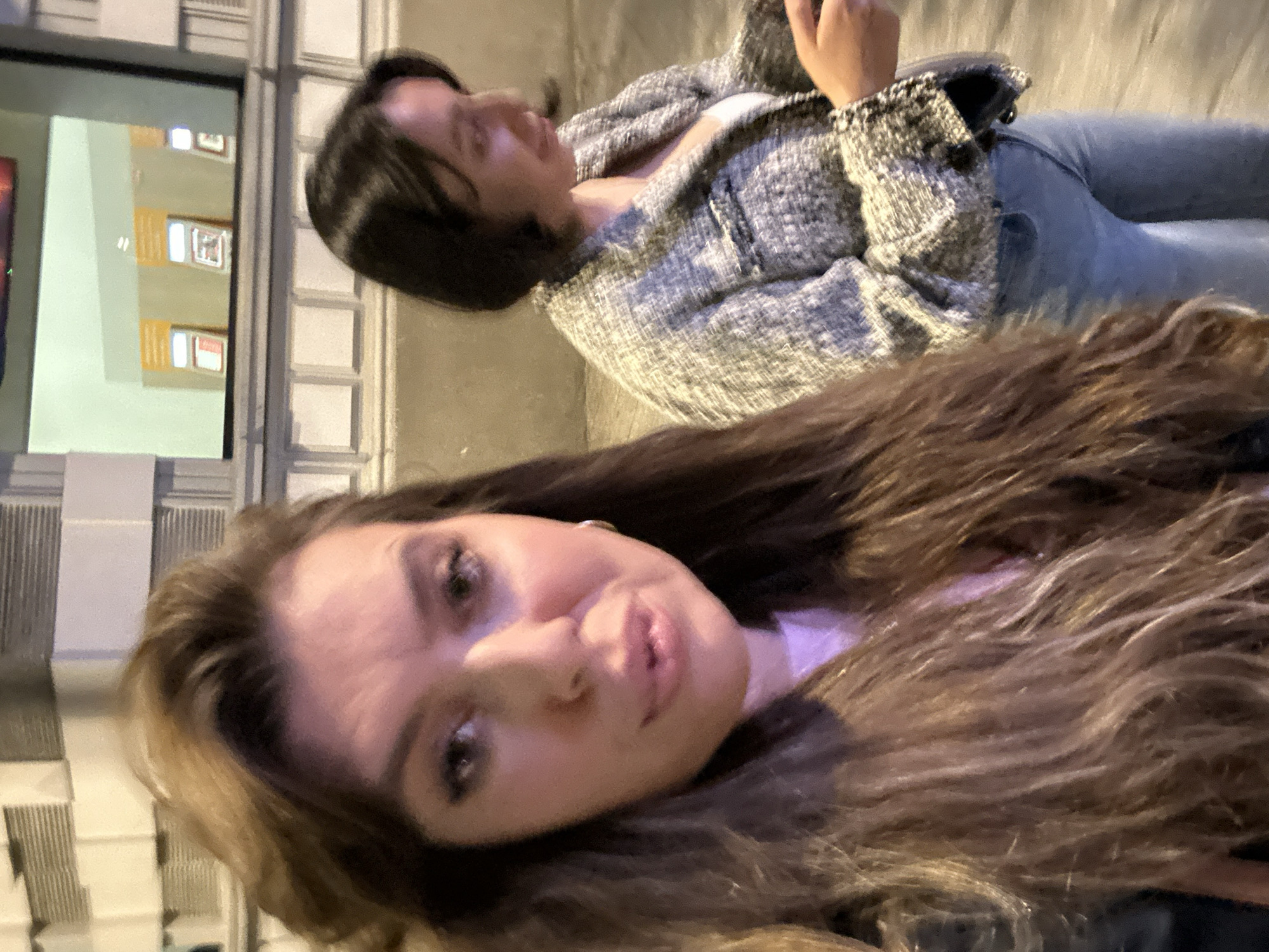 The image shows two women posing for a photo at night in Batumi, Georgia. One woman is in the foreground with long, brown, wavy hair, light skin, and a neutral expression. The other woman is slightly behind, wearing a tweed jacket and jeans, with short dark hair. A building is visible with a window reflecting lights from inside. The background appears to be a street or sidewalk, possibly with architectural details.