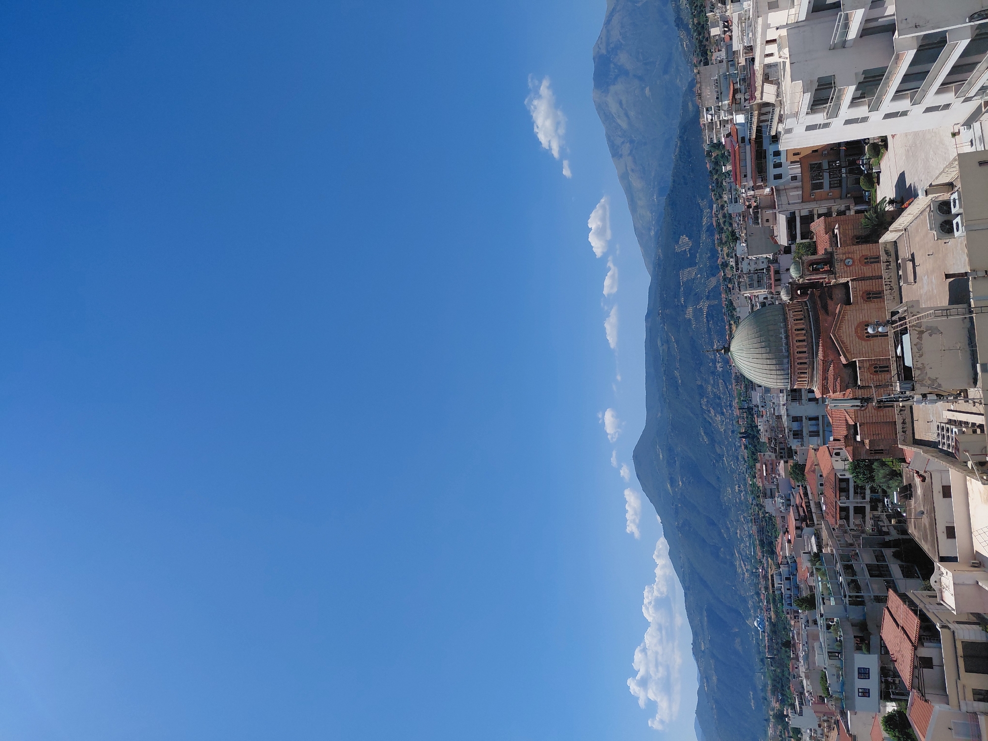 This is a landscape photograph taken from an elevated viewpoint, looking over the city of Aegio, Greece. The sky is a clear, bright blue with a few scattered white clouds. In the mid-ground, a domed building, likely a church or cathedral, is prominent. The building has a reddish-brown facade and a green dome. Surrounding this central structure is a dense urban landscape characterized by multi-story buildings with terracotta roofs. The architecture suggests a Mediterranean style. The background is dominated by a series of rolling hills and mountains, some of which are covered in vegetation. The overall impression is of a sunny day in a coastal or near-coastal Greek city, with historical and natural elements forming the panorama. There are no people visible in the frame.