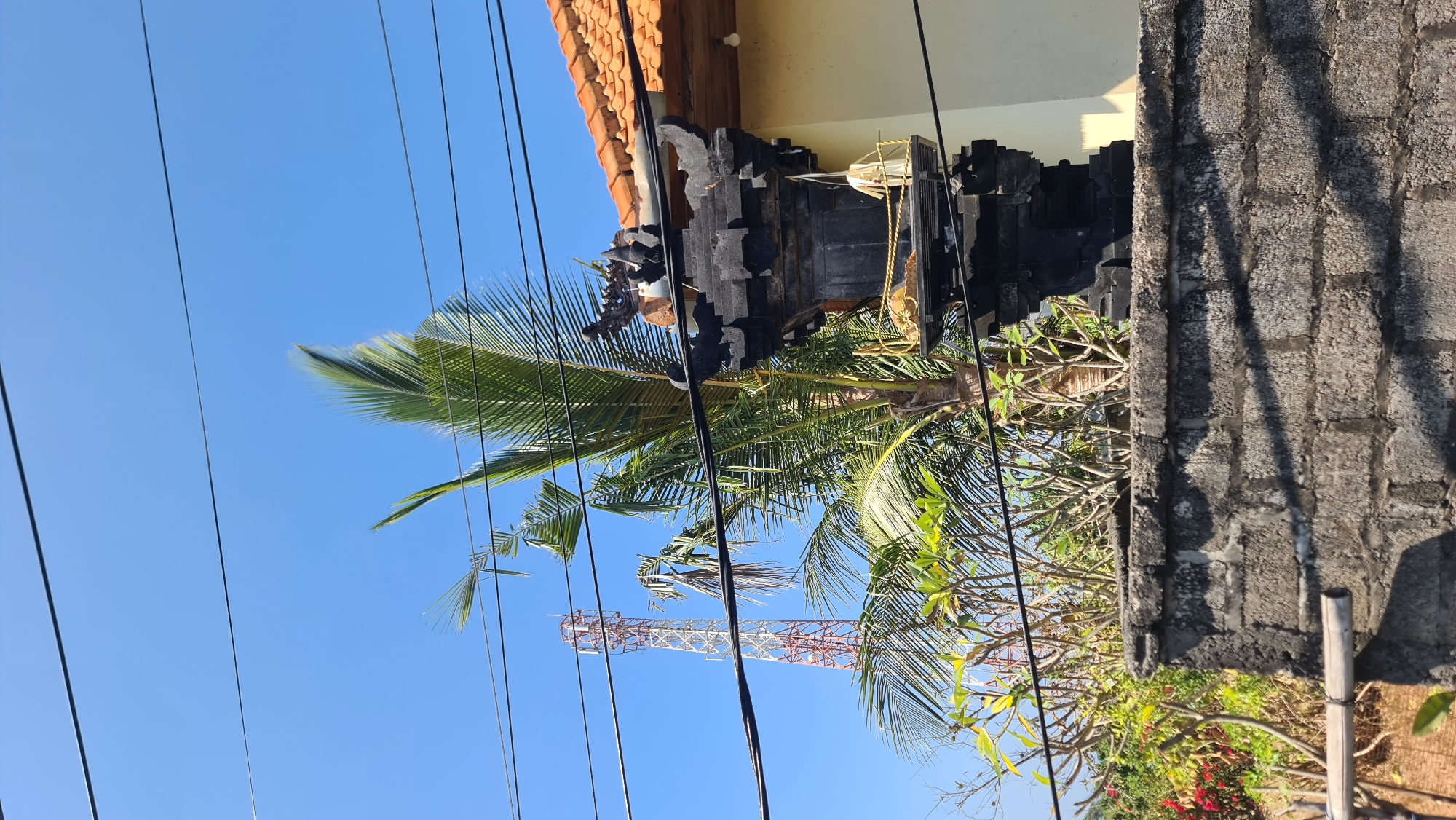 In the city of Kuta Selatan, Indonesia, a clear blue sky serves as the backdrop for a scene dominated by a network of overhead wires. To the left, a tall, red and white lattice tower, likely a telecommunications tower, rises against the horizon. In the foreground, a black stone Balinese temple structure, intricately carved, stands atop a textured concrete wall. Palm fronds from a nearby tree are visible, as are smaller, leafy plants. To the right, the edge of a building with an orange tiled roof is partially visible. The overall impression is one of a tropical, urban environment with a blend of modern infrastructure and traditional architecture.