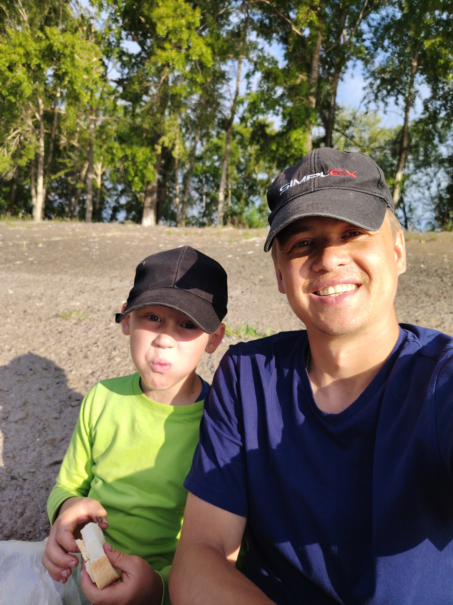 In Chaikovsky, Russia, two people, a man and a child, pose for a selfie outdoors. The man, wearing a dark blue t-shirt and a black baseball cap with "Simplex" embroidered on it, smiles at the camera. Beside him, a young boy in a bright green long-sleeved shirt and a black baseball cap makes a puckered face. The boy holds a piece of white bread in his hands. They are seated on a gravelly surface, with a backdrop of trees and a hint of blue sky. The lighting suggests it is daytime, possibly late afternoon, given the long shadows.