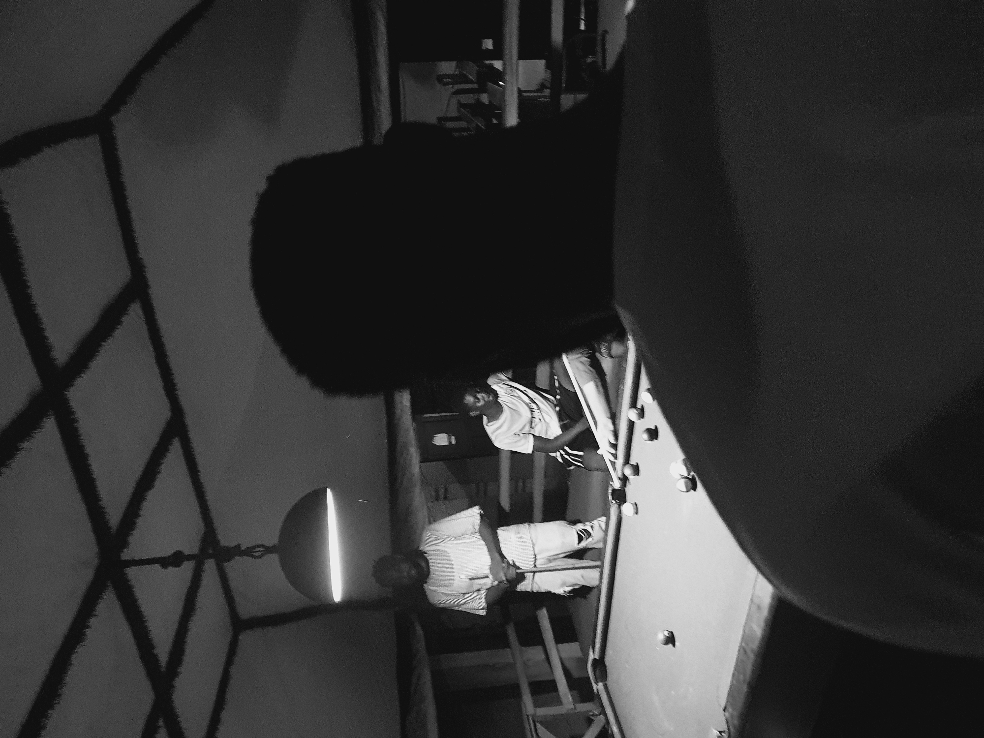 The image depicts an indoor pool hall scene in Lokoja, Nigeria. Three individuals are present. The foreground is dominated by the silhouette of a person facing away from the camera, obscuring much of the view. In the mid-ground, two men are actively engaged in a game of pool. One man, standing, is holding a pool cue and appears to be taking a shot, while the other man is in a crouched position, also with a cue, ready to play. The pool table itself is visible, with several billiard balls scattered across its surface. The environment suggests a somewhat dimly lit, casual establishment, with a hanging light fixture providing illumination and a wooden ceiling structure overhead. The overall mood is one of leisure and friendly competition.