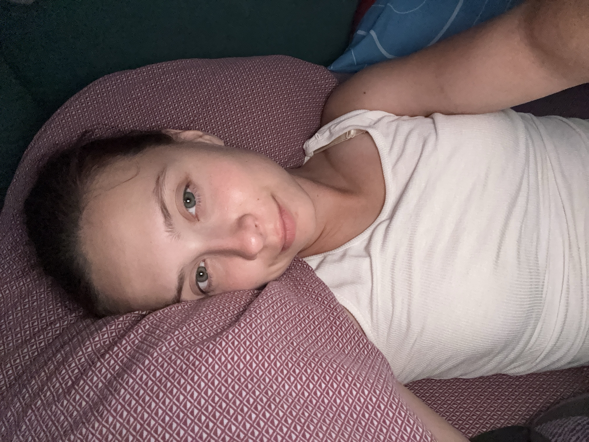 A young woman with light skin, green eyes, and light brown hair is lying down and looking at the camera. She is wearing a white tank top. She is resting her head on a purple pillow with a diamond pattern. Behind her is a dark green surface and a blue pillow with white lines. The scene appears to be indoors, likely in a bedroom. There is no identifiable information about the location in the city of Прикамский, Russia.