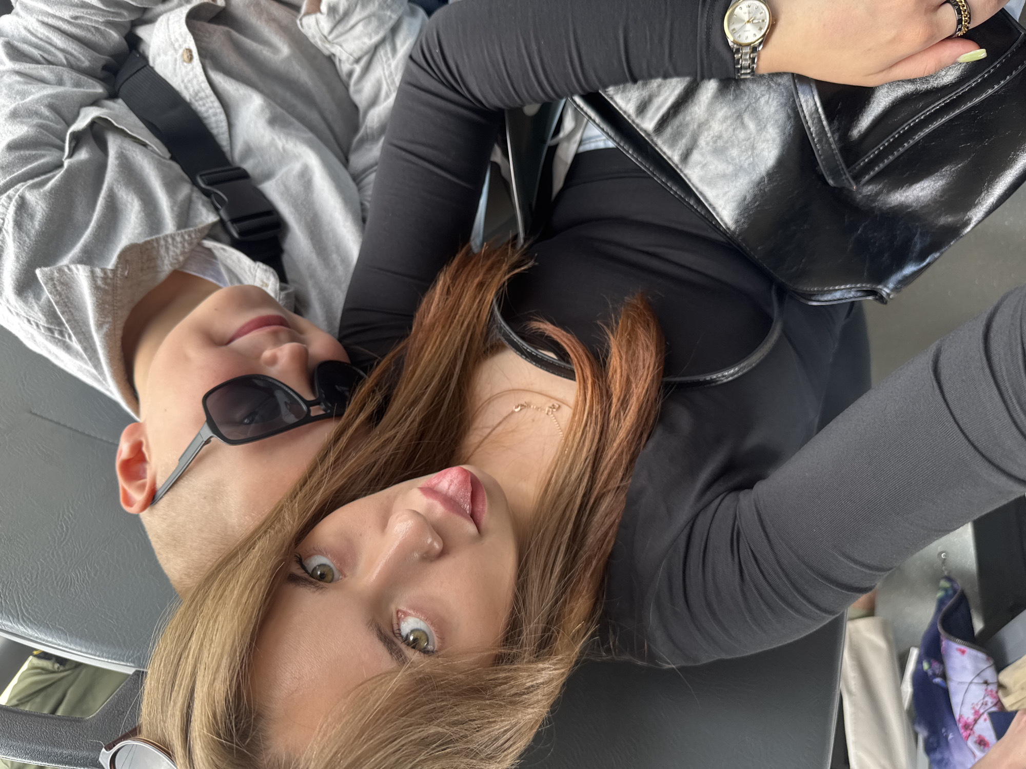 A young woman and a boy are taking a selfie on a bus in Chaikovsky, Russia. The woman is in the foreground, sticking her tongue out at the camera. She has long, light brown hair with reddish-brown ends and is wearing a black long-sleeved shirt and a black purse. She also has a watch on her left wrist and rings on her fingers. The boy is to her right, wearing sunglasses and a light gray collared shirt. He is also wearing a black strap across his chest. The background shows a gray bus seat and other passengers. The lighting is natural and appears to be daytime. The image is taken from a slightly high angle, looking down at the subjects.