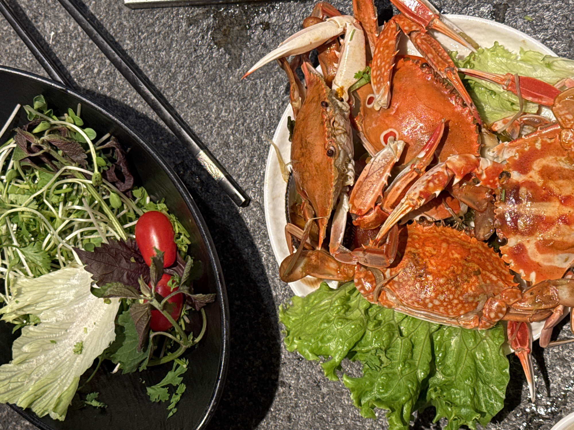 The image shows a plate of cooked crabs surrounded by lettuce, and a black bowl containing various greens and cherry tomatoes. Chopsticks are visible next to the bowl. The scene is likely a restaurant setting, possibly in Đà Nẵng, Vietnam, though this is speculative and based on the type of seafood dish. No people are visible in the image.