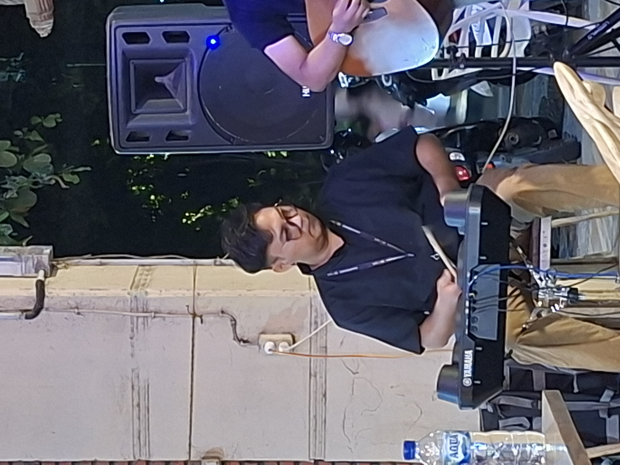 A medium shot depicts a man playing a black Yamaha electronic drum kit. He wears glasses and a black shirt with a lanyard around his neck. The location is Lhokseumawe, Indonesia, suggested by the presence of an "AQUA" branded water bottle. Above him, partially obscured, is another person playing an instrument. A large black speaker is positioned to the left, and sections of a painted white wall are visible behind the subject.
