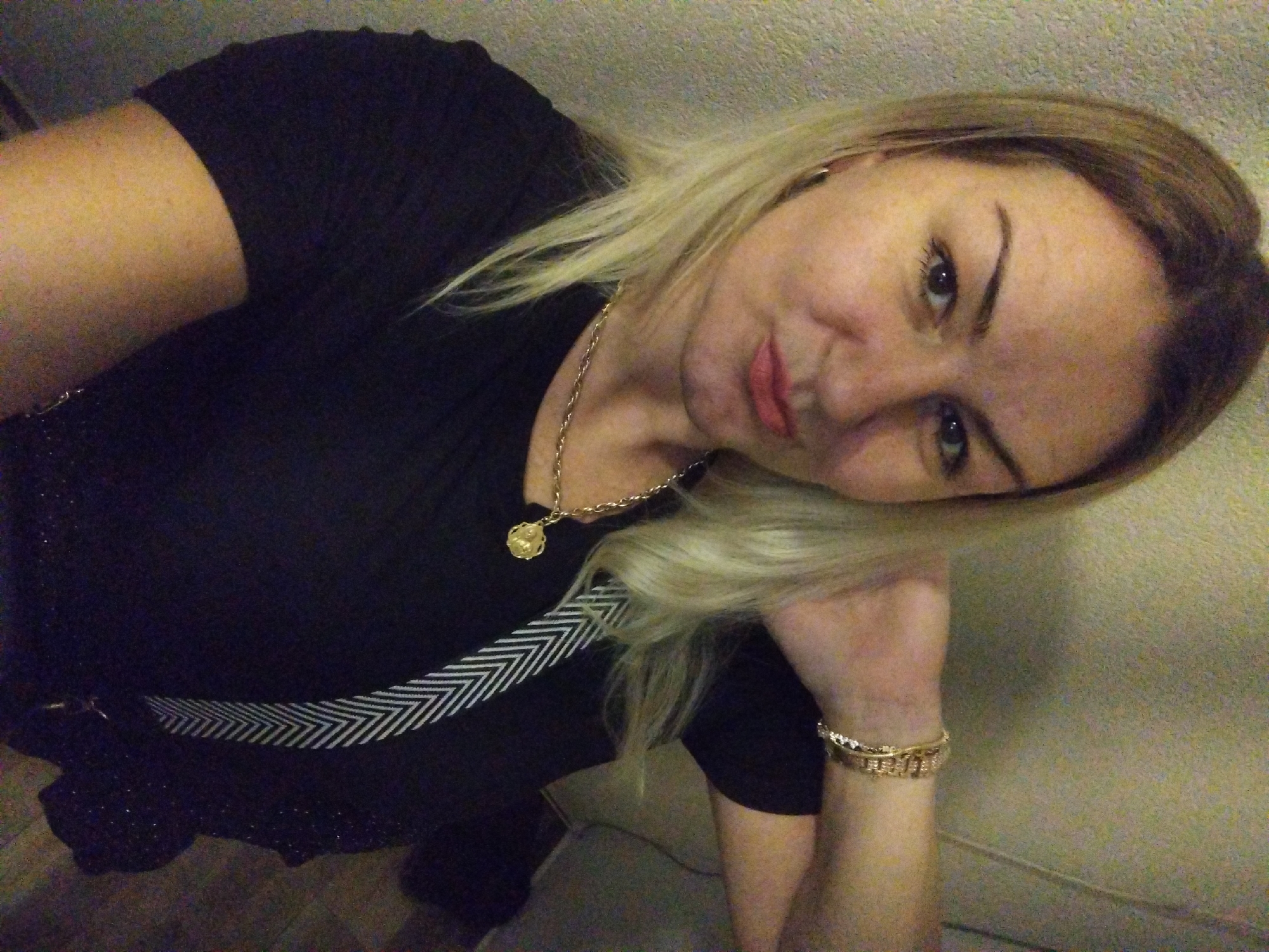 A woman with blonde hair and fair skin is posing for a selfie indoors. She is wearing a black t-shirt with a white chevron pattern on the left shoulder and a gold necklace with a circular pendant. She also wears a gold bracelet on her left wrist, which is resting near a beige or light-colored wall. The setting appears to be a dimly lit room. There is no indication of Stara Zagora, Bulgaria.