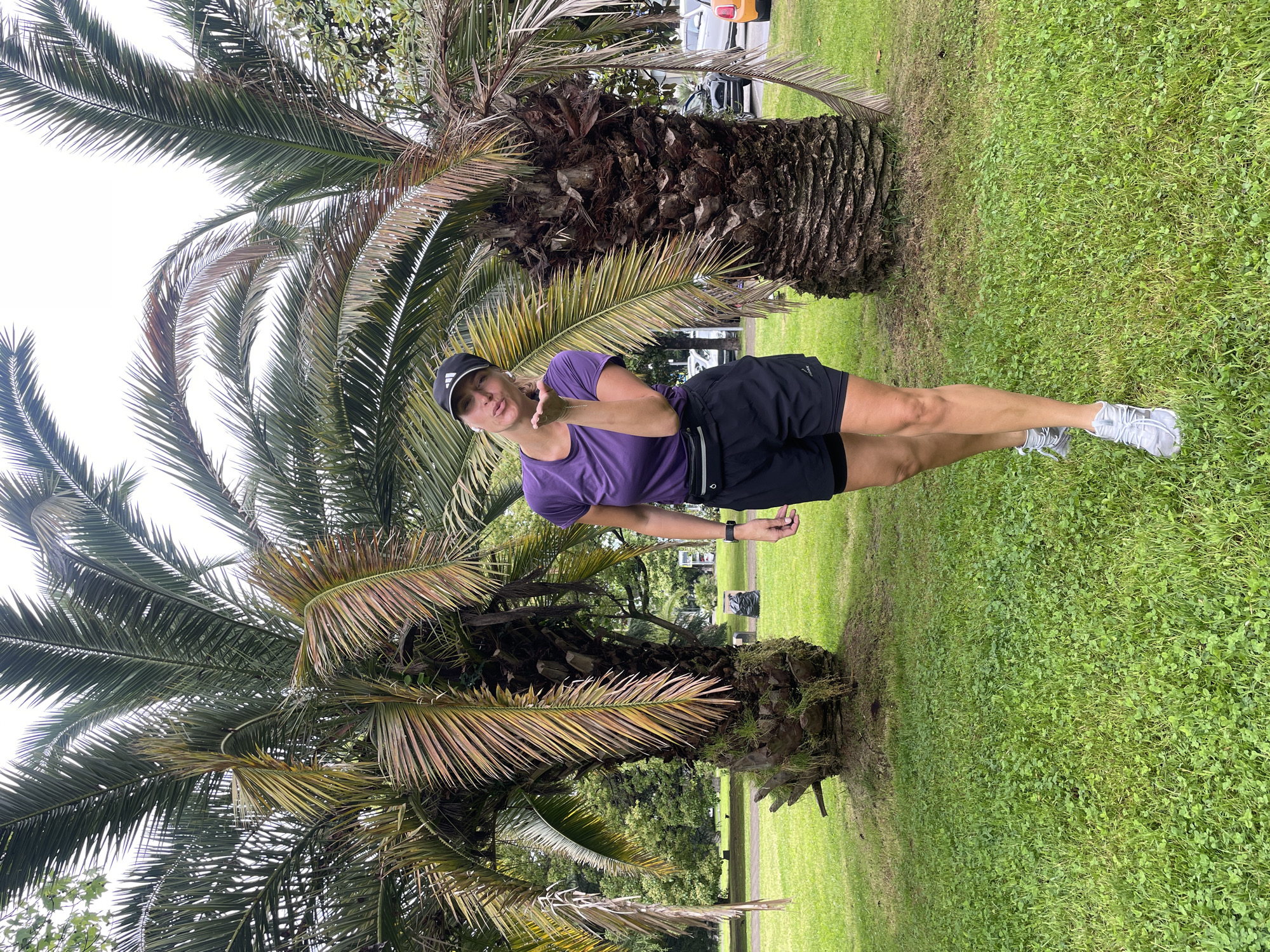 A woman stands in front of two palm trees on a grassy lawn, likely in a park or public space. She is wearing a purple t-shirt, black shorts, a black Adidas baseball cap, and grey running shoes. She is positioned between the two trees with one foot extended forward. She is making a kissing face at the camera and appears to be posing for a photo. The sky is overcast. Other trees are visible in the background. Based on the presence of palm trees, the location is a warm, temperate climate, like Sochi, Russia.