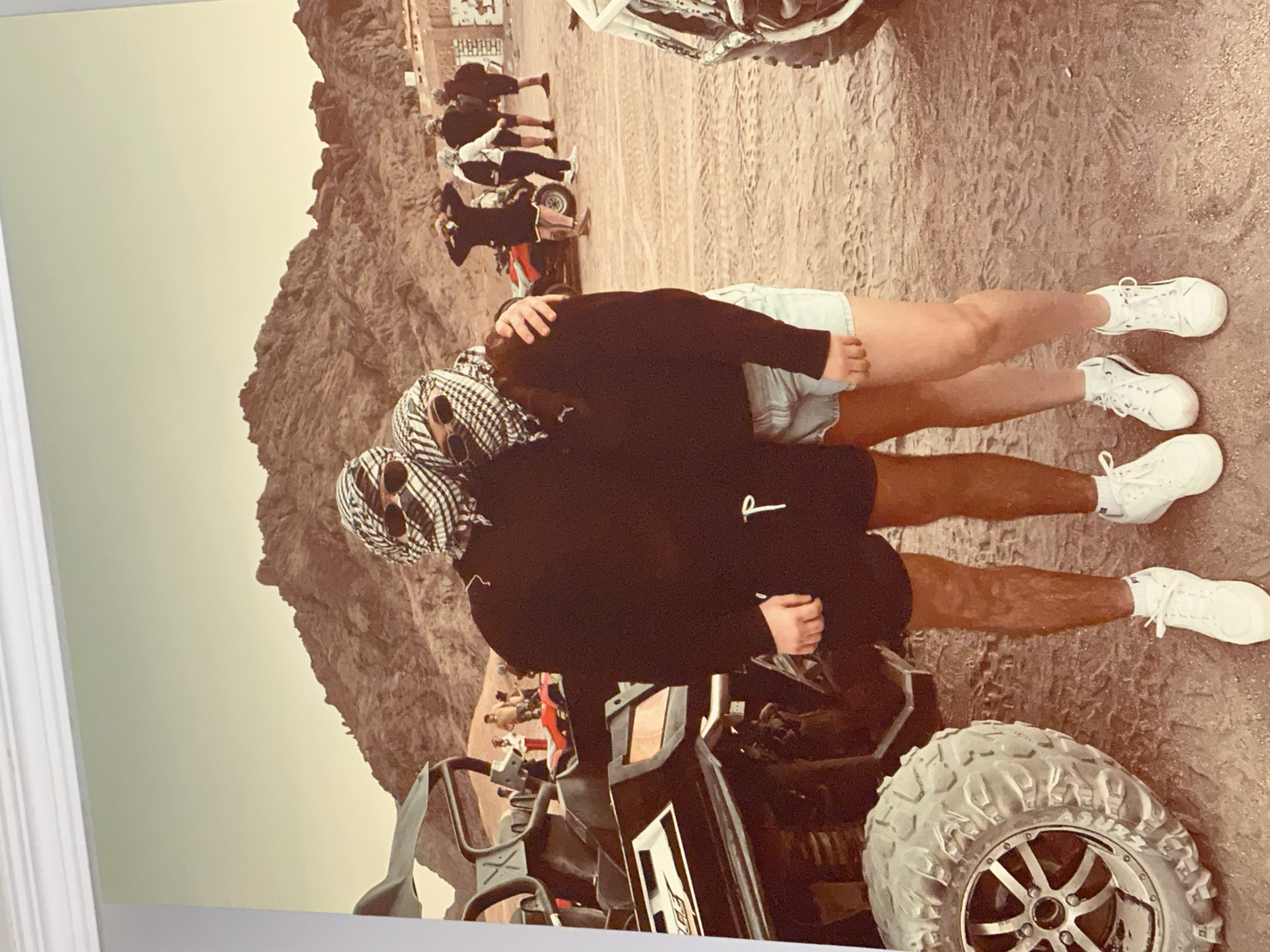 In a desert landscape with rocky mountains in the background, two individuals stand embracing, their faces covered by keffiyeh scarves and wearing sunglasses. The person on the left is wearing black shorts and a black hoodie, while the person on the right is wearing denim shorts and a black hoodie. They are both wearing white sneakers. To their left, a dark-colored ATV is parked. Further in the background, several other ATVs and a group of people are visible. The ground is sandy and appears to be a dirt track. The sky is a pale, overcast yellow.
