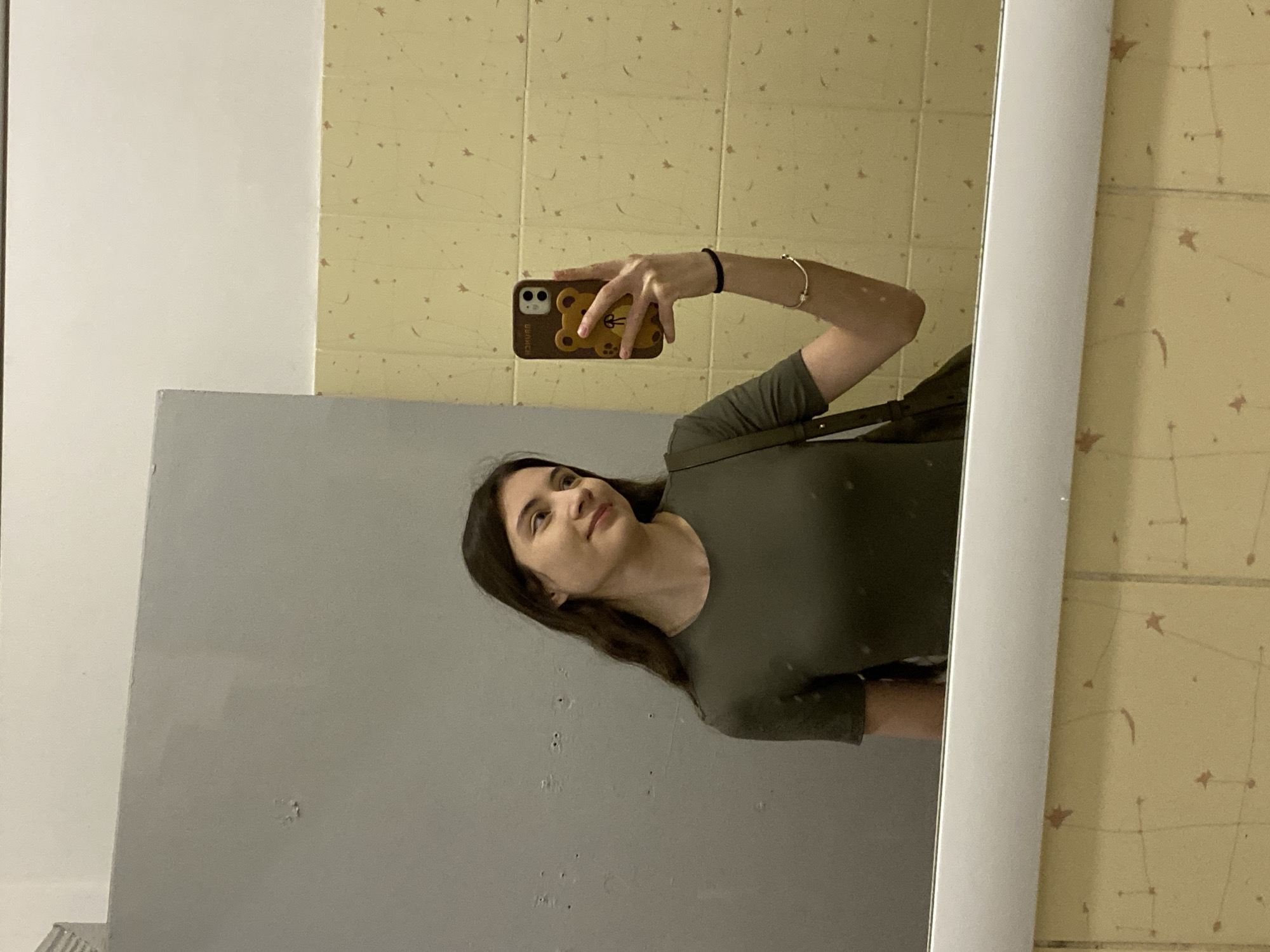 A young woman with long brown hair is taking a selfie in a mirror. She is wearing an olive green t-shirt and holding a smartphone with a brown bear-shaped case. The background reveals beige-colored walls patterned with small star-like shapes, and a grey wall is reflected in the mirror. The location in Votkinsk, Russia, cannot be determined from the visual information provided.