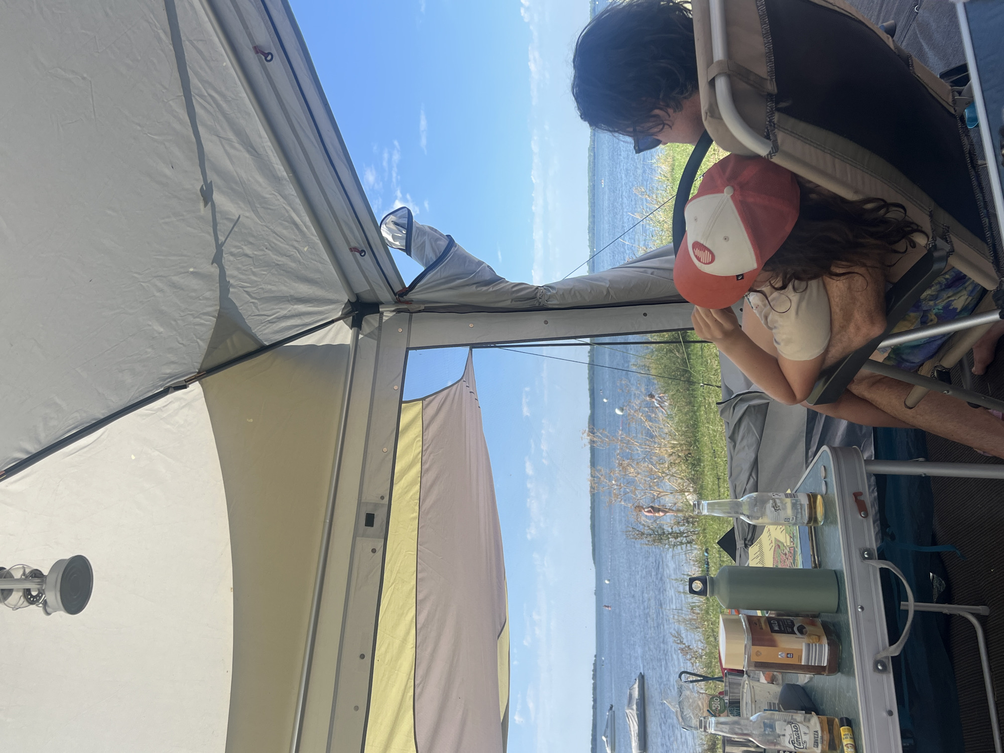 The image shows a campsite by the sea in the city of Am Salzhaff, Germany. In the foreground, a table is set up with two bottles of beer, a jar of coffee, a green water bottle, and other items. Two people are seated in chairs, facing away from the camera, looking out at the water. One person is wearing a red and white baseball cap. The background shows a body of water with boats and a distant shoreline. The sky is blue with some clouds. The overall scene depicts a relaxed outdoor setting.