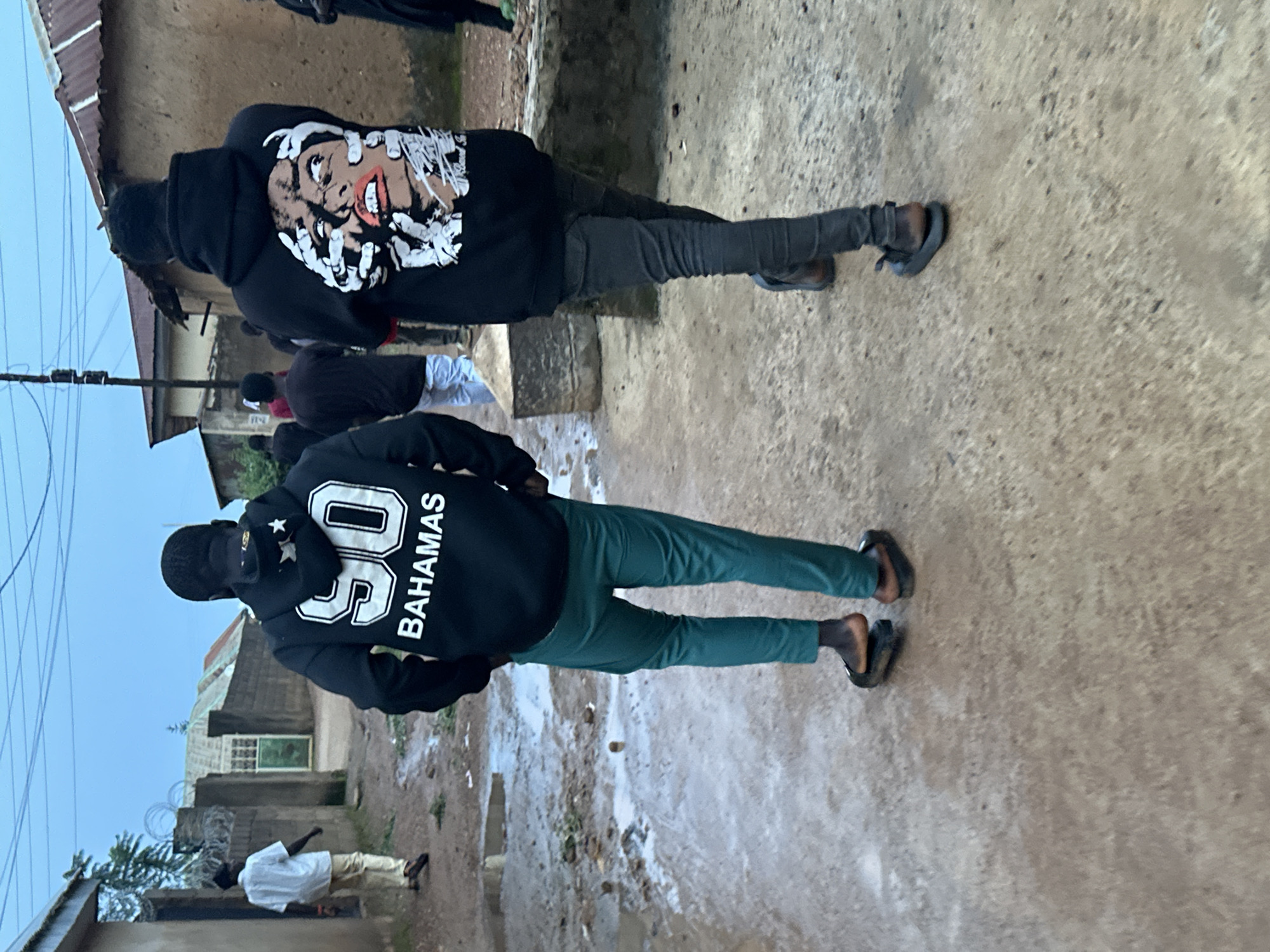 In Du, Nigeria, two young men walk away from the camera down a wet, dirt path lined with buildings. The man on the left wears a black hoodie with "90 BAHAMAS" printed on the back in white, and teal-colored pants. He is wearing flip-flops. The man on the right wears a black hoodie with a graphic of a face being grabbed by hands on the back, and dark jeans. He is wearing dark-colored sneakers. The sky is overcast and the ground is muddy and wet, with puddles visible. In the background, other people can be seen walking, and buildings with corrugated metal roofs are present.