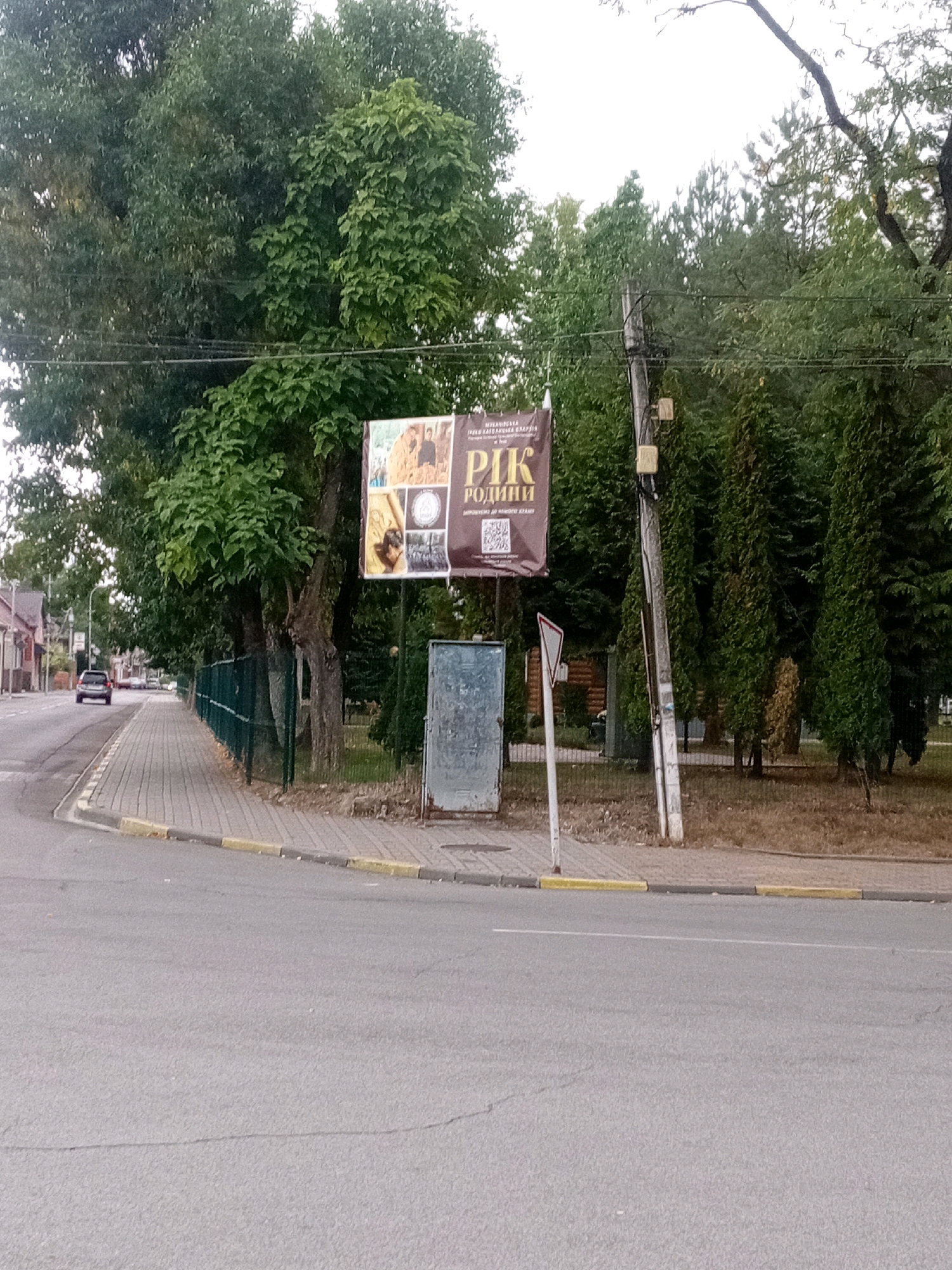 The image captures a street scene in Tiachiv, Ukraine. A gray asphalt road curves to the right, bordered by a paved sidewalk with yellow and gray curbs. A dark-colored car is visible on the road, driving away from the camera. Along the sidewalk, a green metal fence runs parallel to the road, separating it from a grassy area with trees. A large banner hangs from a utility pole on the right side of the frame, featuring text and images, including what appears to be a QR code. A metal cabinet and a triangular road sign are also present near the utility pole. The background consists of lush green trees and foliage, with some buildings visible in the distance on the left side of the image. The sky is overcast.