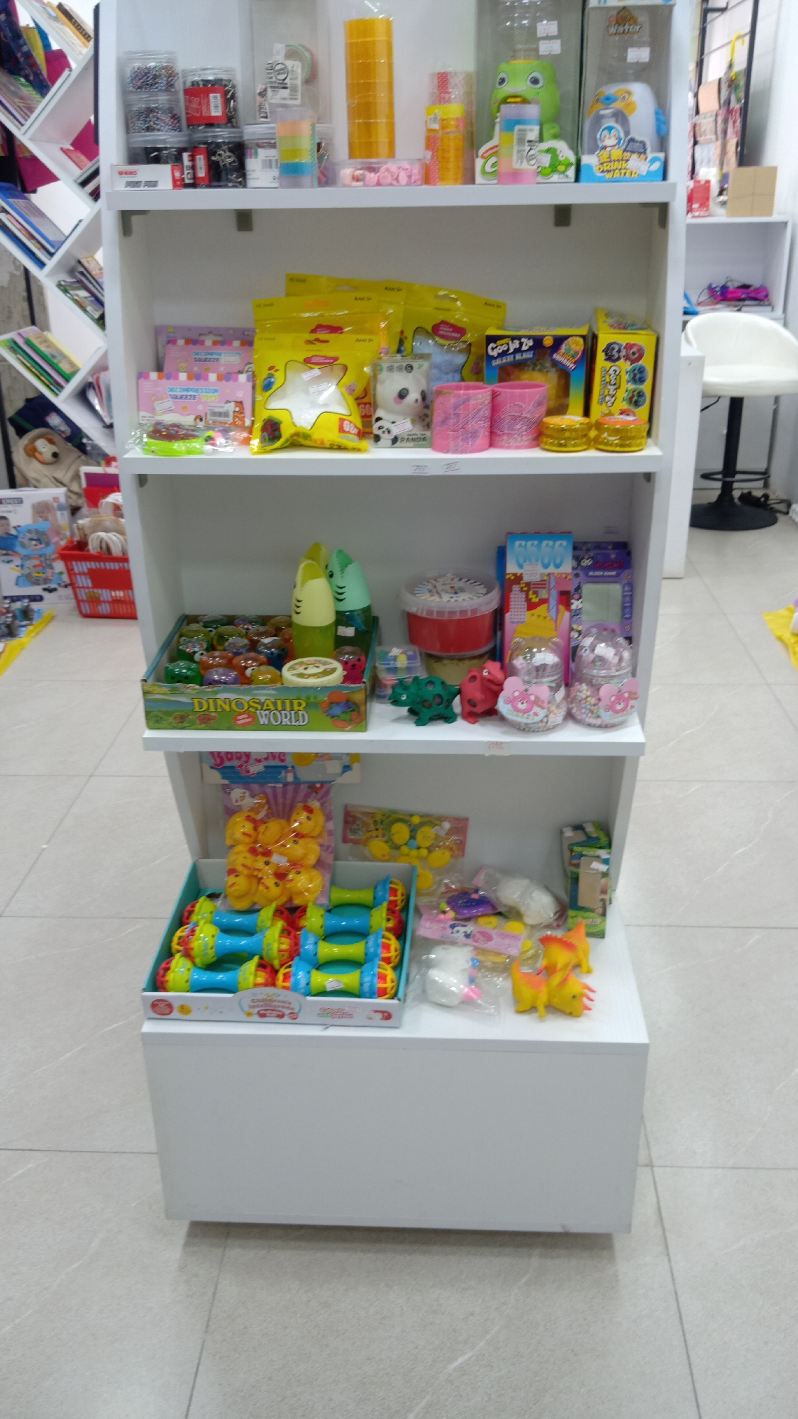 A three-tiered white shelving unit is filled with various toys and stationery items, likely in a retail setting. The top shelf contains containers of beads, rolls of tape, and water dispenser toys. The second shelf features bagged and boxed toys, containers of putty, and a "Dinosaur World" box filled with small toys. The bottom shelf holds more toys, including baby rattles and dinosaur figures. The floor is tiled. In the background, there are bookshelves and a white chair, suggesting a store environment.